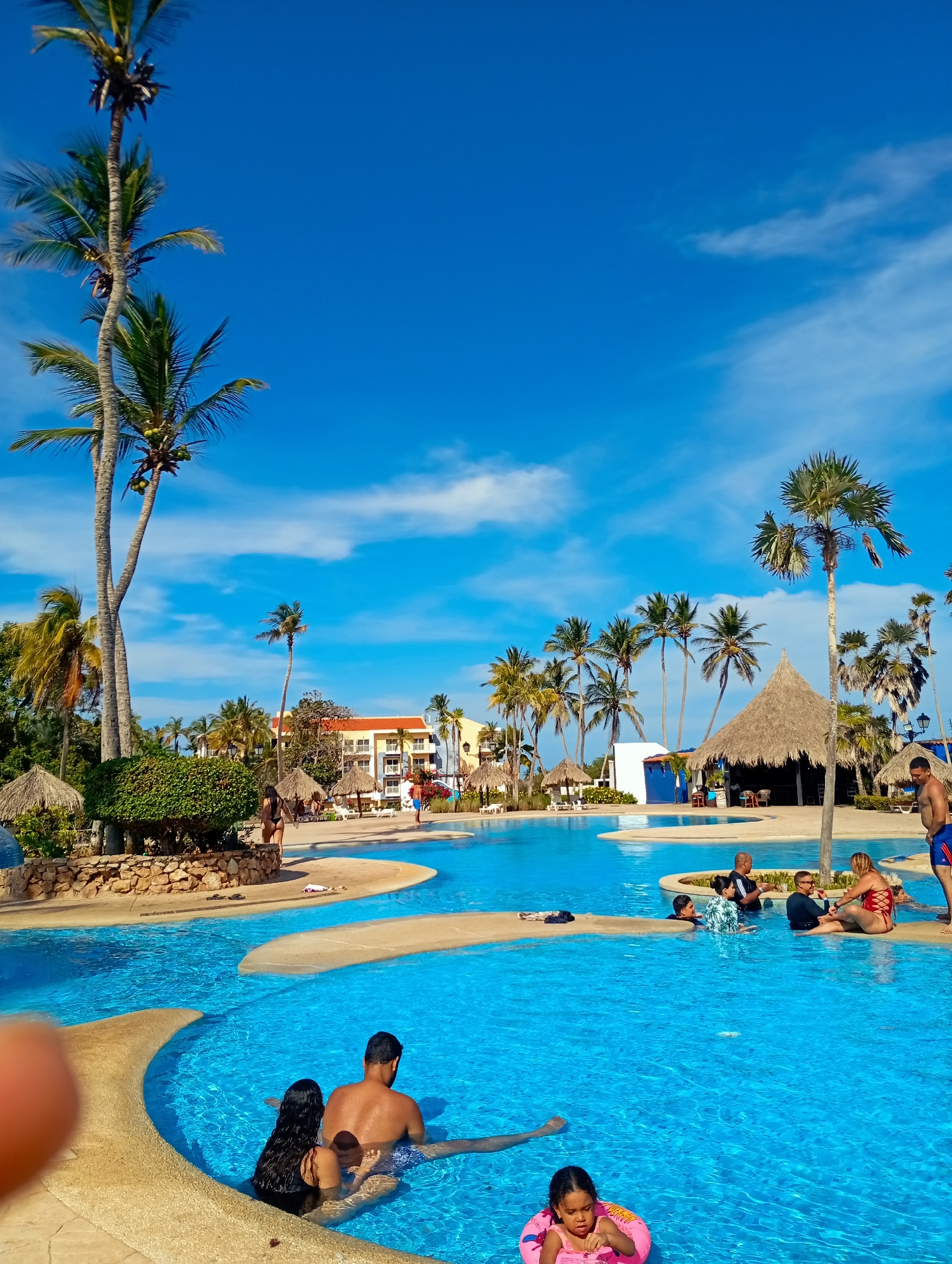 This image depicts a vibrant, sunny tropical resort scene at Playa El Agua, Venezuela. The central focus is a large, multi-level swimming pool with clear, sparkling blue water, surrounded by beige-colored decking.
Numerous people are enjoying the pool area. In the foreground, a man and a woman are partially submerged in the water, appearing to relax. Further back, a young child in a pink inflatable swimming ring is visible. Other adults are scattered throughout the pool, some standing, some seated, and others conversing.
The setting is lush and tropical, characterized by many tall palm trees, some bearing green coconuts, reaching into a brilliant clear blue sky dotted with sparse white clouds. This suggests a bright, sunny day, likely midday or early afternoon. Various straw-roofed huts or palapas are positioned around the pool and grounds, providing shade and adding to the resort ambiance. One larger palapa in the mid-ground appears to be a bar or seating area.
In the background, a multi-story hotel building with yellow and orange sections is visible, surrounded by more greenery and palm trees. The overall activity involves leisurely swimming, relaxing, and socializing by the pool. The scene conveys a sense of vacation, warmth, and enjoyment. There is no visible text in the image.