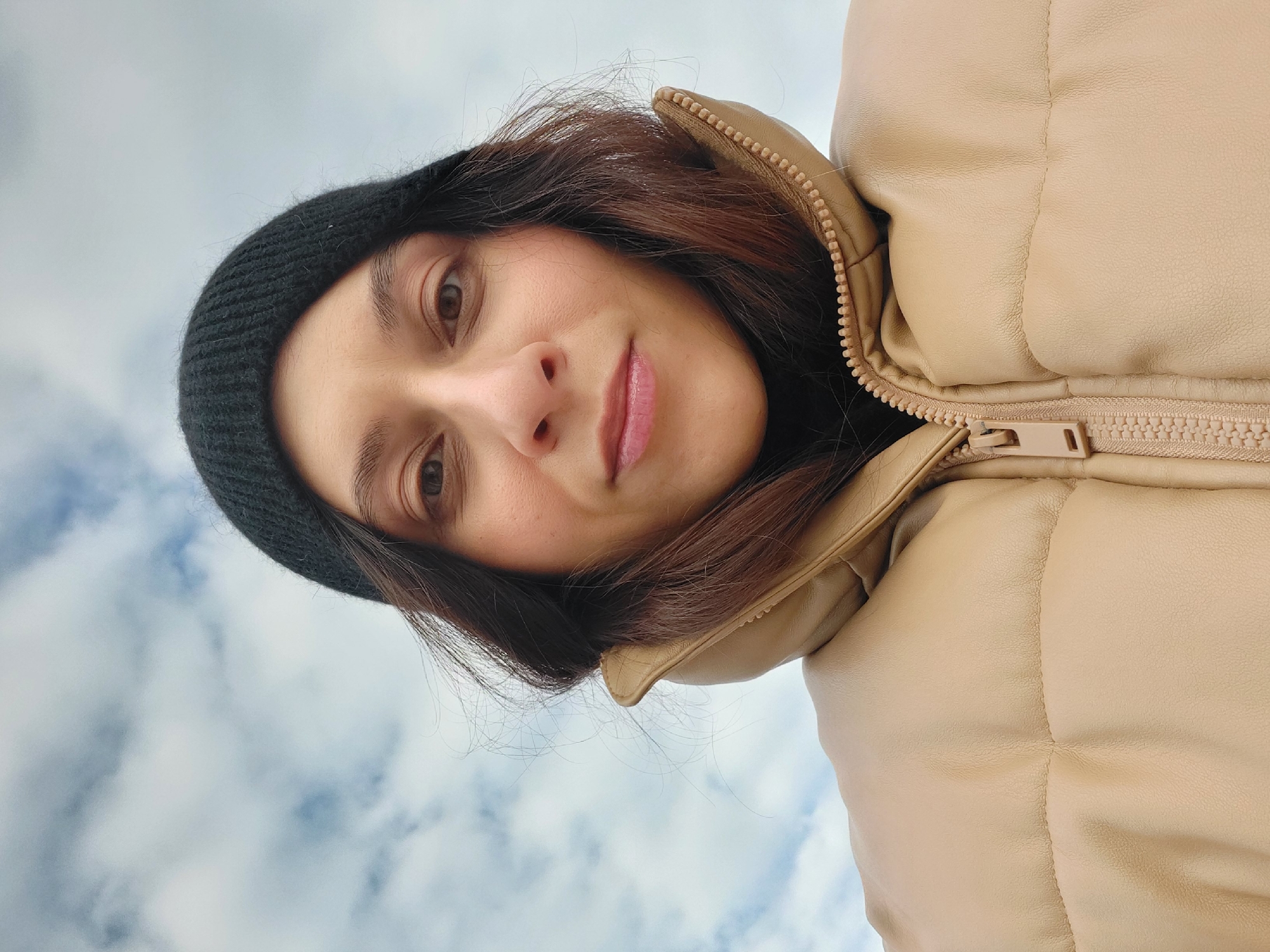 A woman with fair skin, dark brown hair, and a black beanie is seen wearing a light beige puffy jacket with the zipper partially visible. She is looking directly at the camera with a slight smile. The background shows a cloudy sky. The location is unknown and there is no information in the image to suggest it is Novosibirsk, Russia.