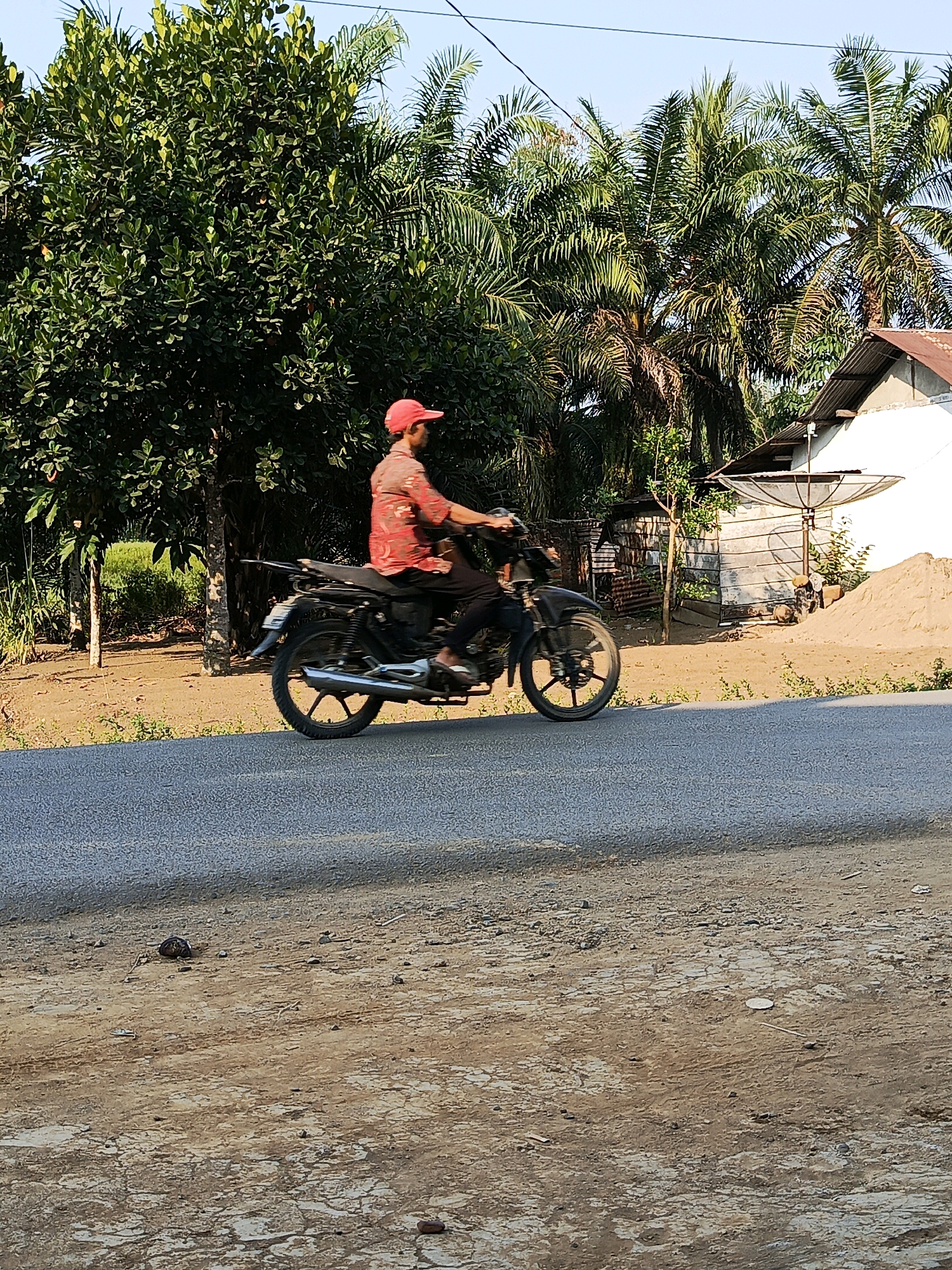 In Megang Sakti I, Indonesia, a man wearing a red cap and a red patterned shirt rides a black motorcycle on a paved road. The road is bordered by dry, dusty ground with sparse vegetation. Behind the road, a dense green tree is visible, with palm trees and a building in the background. The building appears to be made of corrugated metal with a rusted metal roof and a satellite dish attached. A pile of sand sits next to the building. The sky is clear and blue.