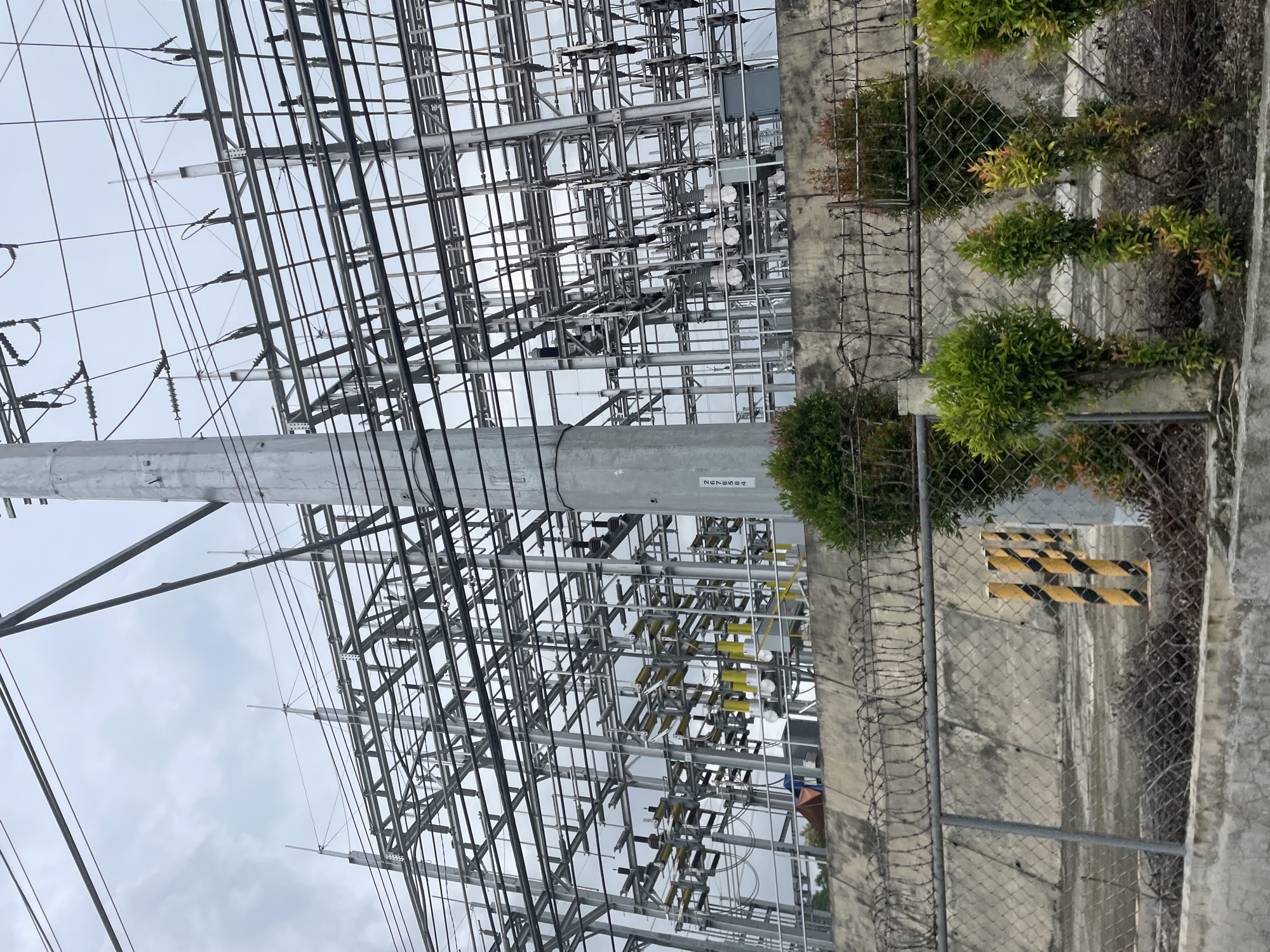 The image depicts an electrical substation in Davao City, Philippines, under an overcast sky. The substation features a complex network of metal structures supporting numerous wires and electrical components, including insulators and what appear to be transformers. A tall, cylindrical concrete pole stands prominently in the foreground, with electrical wires extending from it and connecting to the substation. A concrete wall forms a boundary, topped with barbed wire fencing. In front of the wall, chain-link fencing is visible, with small, manicured green shrubs planted along its base. Several yellow and black striped bollards are positioned behind the chain-link fence. The overall scene is utilitarian, typical of industrial infrastructure.