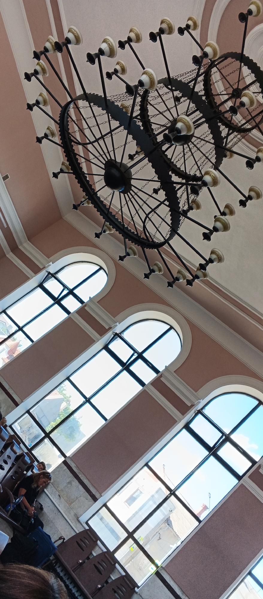 The image depicts an interior view of what appears to be a grand hall or waiting area with a distinctive chandelier and arched windows. The chandelier is elaborate, made of dark metal with multiple lights featuring white and gold accents. The ceiling is a pale pink with decorative white trim.
Below the ceiling, three large, arched windows dominate the scene, angled upwards and inwards. These windows are divided into panes by dark frames and look out onto an urban environment with buildings and a bright sky, suggesting it is daytime. The light filtering through the windows illuminates the interior.
In the foreground, towards the bottom left of the image, there are rows of dark brown chairs, some of which are occupied by people. A woman with blonde hair, wearing a black top, is seated, looking down at something in her lap. Bags, including a blue suitcase, are placed near the chairs. The walls are a muted pink color with decorative horizontal trim.
The image appears to be taken from a low angle, looking upwards towards the chandelier and windows. Based on the architectural style and the presence of what seem to be public waiting areas, this could be a train station, a historical building, or a public institution. A search of Mukachevo, Ukraine, reveals that the Mukachevo railway station has a similar aesthetic, featuring arched windows and decorative elements, making it a plausible location for this scene.