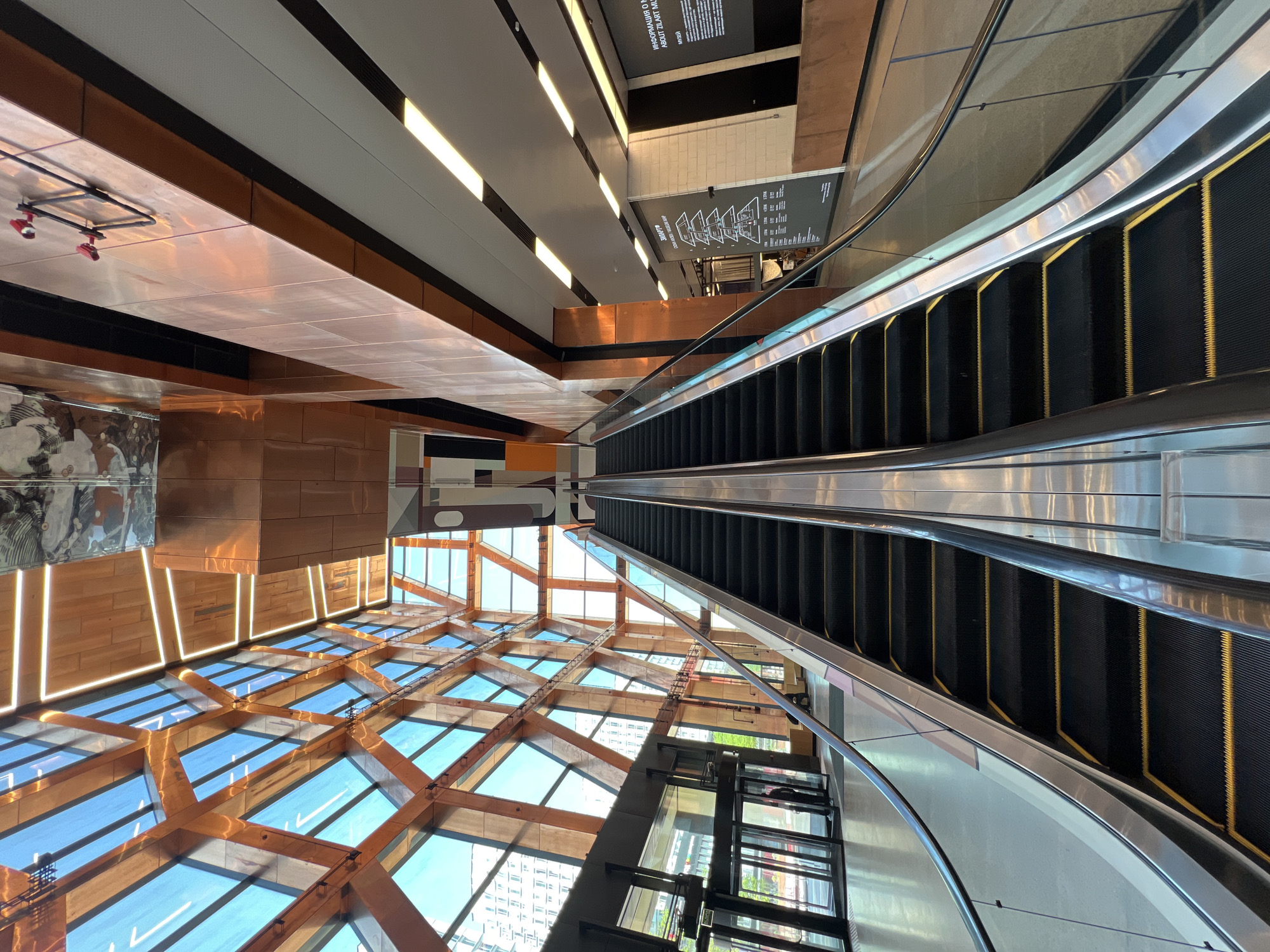 The image captures the interior of the Zaryadye Park Media Center in Moscow, Russia, from a high vantage point, likely within the building itself. A prominent feature is a long escalator leading downwards. To the left of the escalator is a glass-enclosed elevator. The building's architecture is characterized by a grid-like pattern of wooden beams and glass panels forming a skylight, allowing ample natural light to enter. Above the skylight, there are copper-colored structures and a white ceiling with linear light fixtures. Signage, including a map or directory, is visible on the wall near the escalator. No people are visible in the immediate scene.