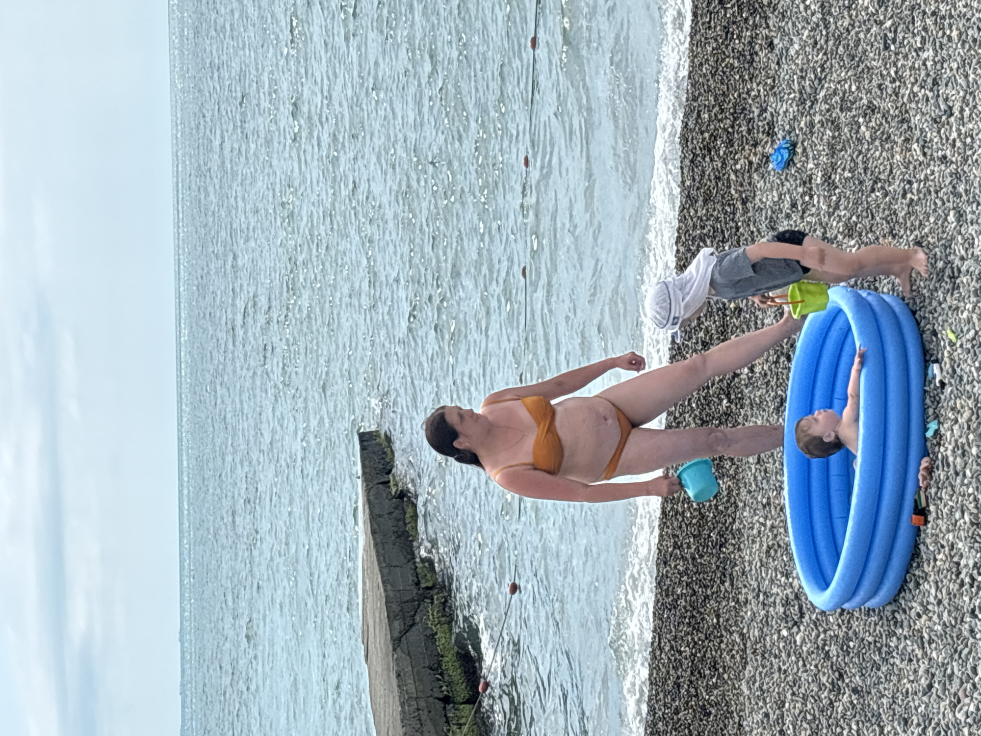 This media file shows a group of people at a beach in Sochi, Russia. The subjects are a woman in an orange bikini and two young children. One child is in an inflatable blue pool on the pebble beach, and another child is standing next to the pool, reaching into a yellow bucket. The woman is standing near the inflatable pool, holding a blue cup.
The scene is a beach with the sea in the background. The water is light blue and appears calm, with small waves gently reaching the shore. The beach itself is covered in grey and brown pebbles. There is a concrete structure in the water to the left of the frame, possibly part of a pier or breakwater. The sky is overcast and a pale blue.
The time of day appears to be daytime, given the lighting. The weather seems to be mild, suitable for beach activities, as indicated by the people in swimwear. The overall mood appears to be relaxed and enjoyable, with the children engaged in play. There is no visible text in the media file.