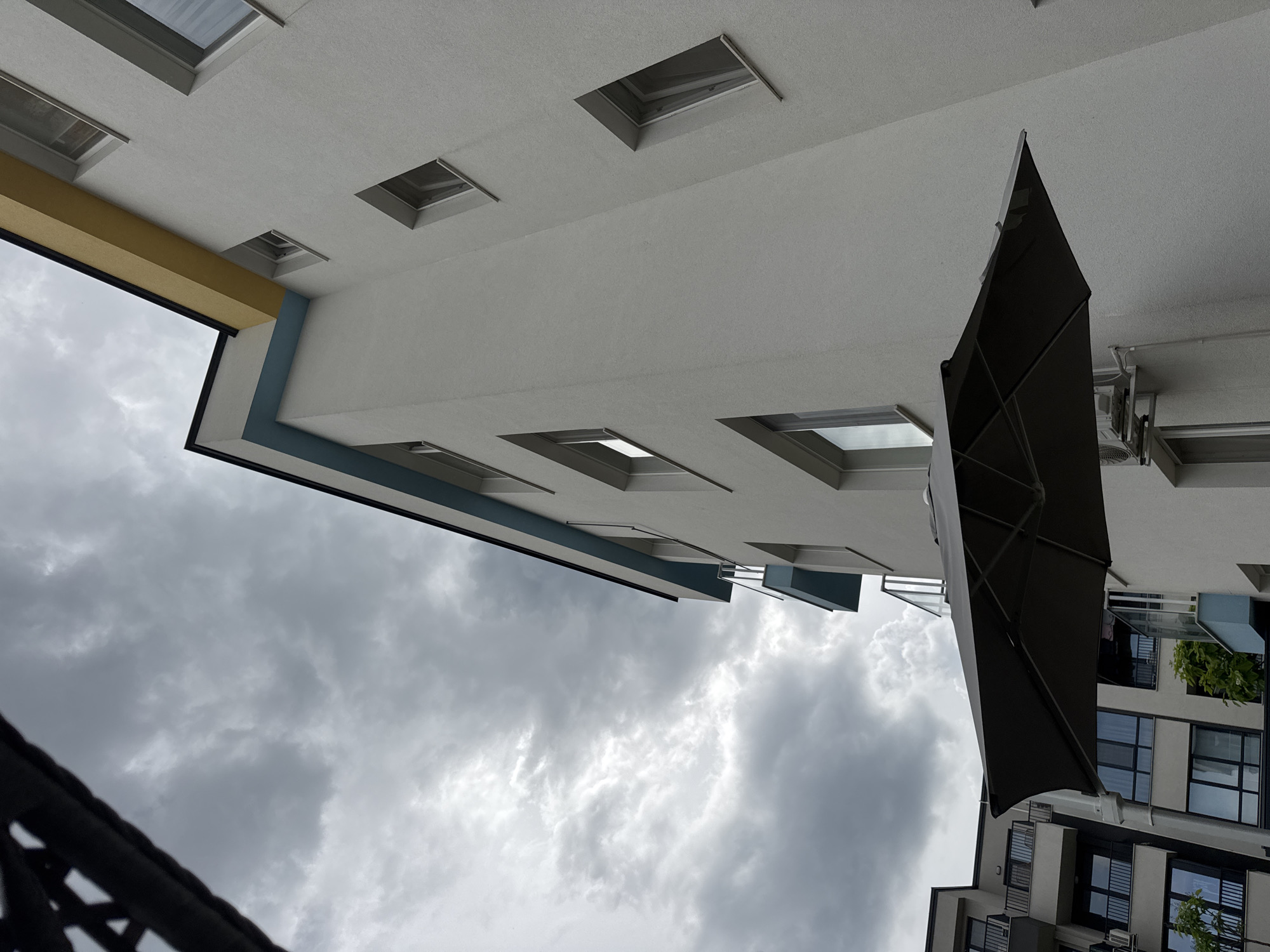 The image shows a view looking up at a building with a cloudy sky in the background. The building has a modern design with white and light gray facades, accented with a yellow band and a blue trim. Several rectangular windows or skylights are visible on the building's exterior, some appearing open. A large, dark umbrella is positioned prominently in the foreground, angled upwards. Beyond the immediate building, another section of the building or a neighboring structure is visible, featuring more windows and balconies. Some greenery can be seen on the balconies. The sky is filled with dark, dramatic clouds, suggesting an overcast or stormy day. The orientation of the image is such that the ground appears to be at the top and the sky at the bottom, indicating the photo was taken from a low vantage point, looking upwards. No people are visible, and no text is discernible in the image. The overall mood is somewhat dramatic due to the stormy sky and the upward-angled perspective.