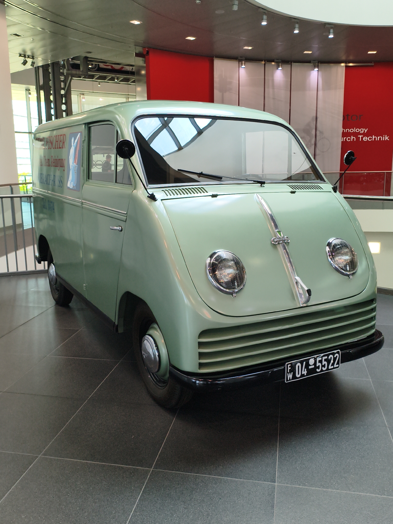 The image shows a vintage light green van parked on a tiled floor in a modern building. The van has a rounded front with two large circular headlights and a chrome strip running down the hood. A license plate with the number "W 04-5522" is mounted on the lower front bumper. The side of the van has text that appears to read "FISCHER Chem. Reinigung" and a phone number "Tel. 6074". In the background, there are large windows, metal railings, and a red wall with white text that reads "ology nology" and "rch Technik". The ceiling has exposed beams and lighting fixtures. The overall scene suggests the van is on display in a museum or exhibition space, likely in Ingolstadt, Germany, given the context of other images from the Audi Museum mobile.