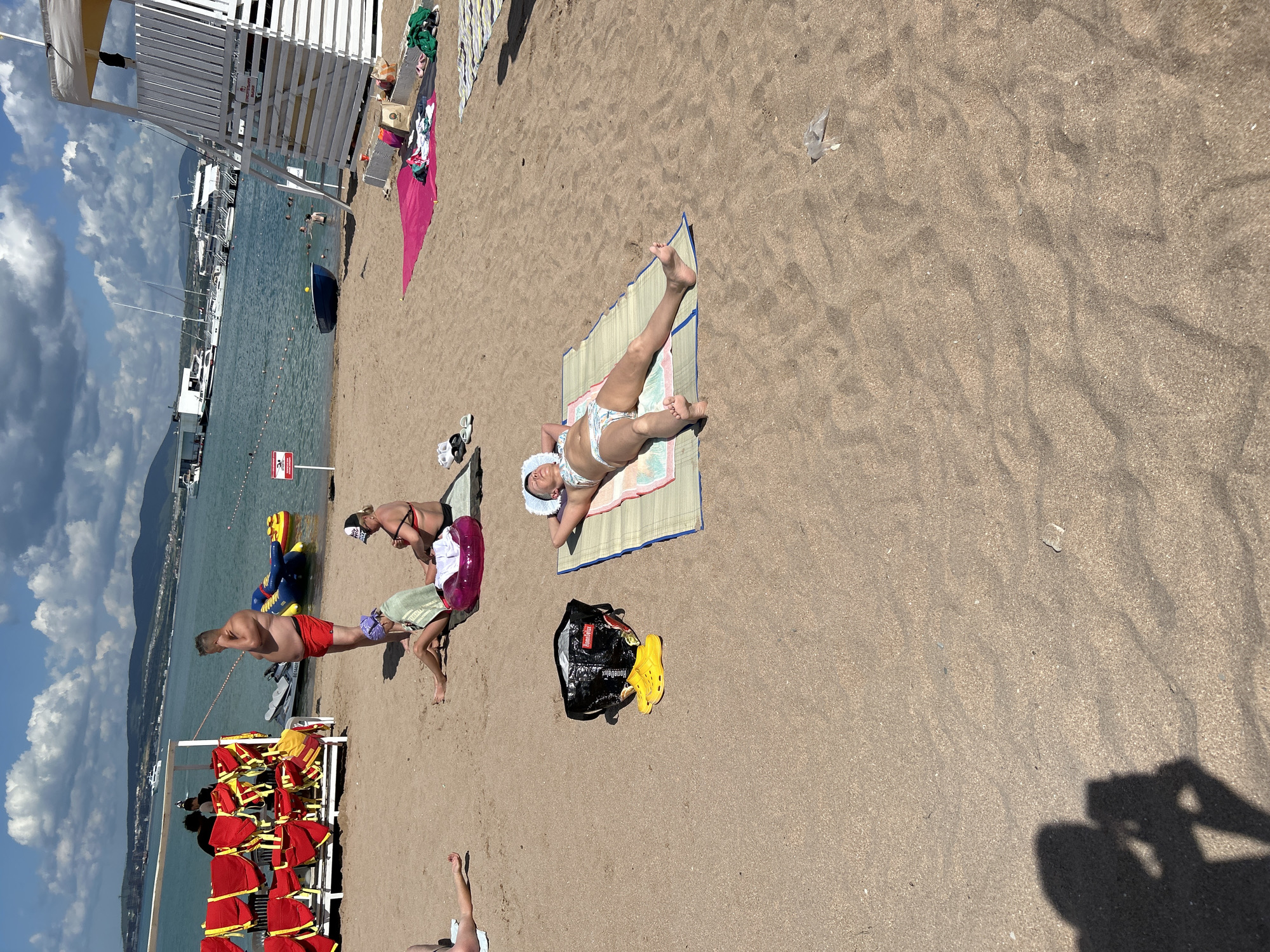 On a sandy beach in Gelendzhik, Russia, under a partly cloudy sky, several individuals are engaged in beach activities. In the foreground, a woman lies on her back on a mat, sunbathing in a patterned bikini. To her left, a black bag and a pair of bright yellow shoes are placed on the sand. Further back, a group of people are seated on towels; one woman wears a bikini and a baseball cap, while another is wrapped in a green towel, and a third is partially visible. In the background, a man in red shorts stands near the water's edge. Lifeguard equipment, including red and yellow vests, is stored on a rack to the left of the frame. The sea is visible, with several boats and yachts anchored offshore. The overall scene depicts a typical summer day at a seaside resort.
