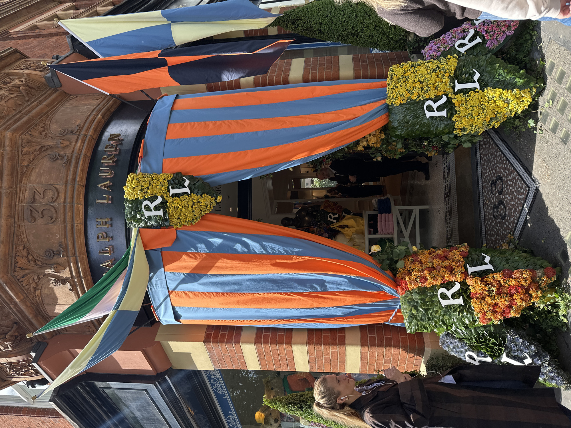 This photo, taken in London, United Kingdom, depicts the exterior of a Ralph Lauren store decorated for an event. The store has striped orange and blue fabric cascading from the top of the entrance and flags in various patterns above. Decorative floral wreaths with the initials "RL" in white are positioned on either side of the entrance. A few people are visible inside the store. A woman wearing a brown coat and white earphones stands in the foreground.