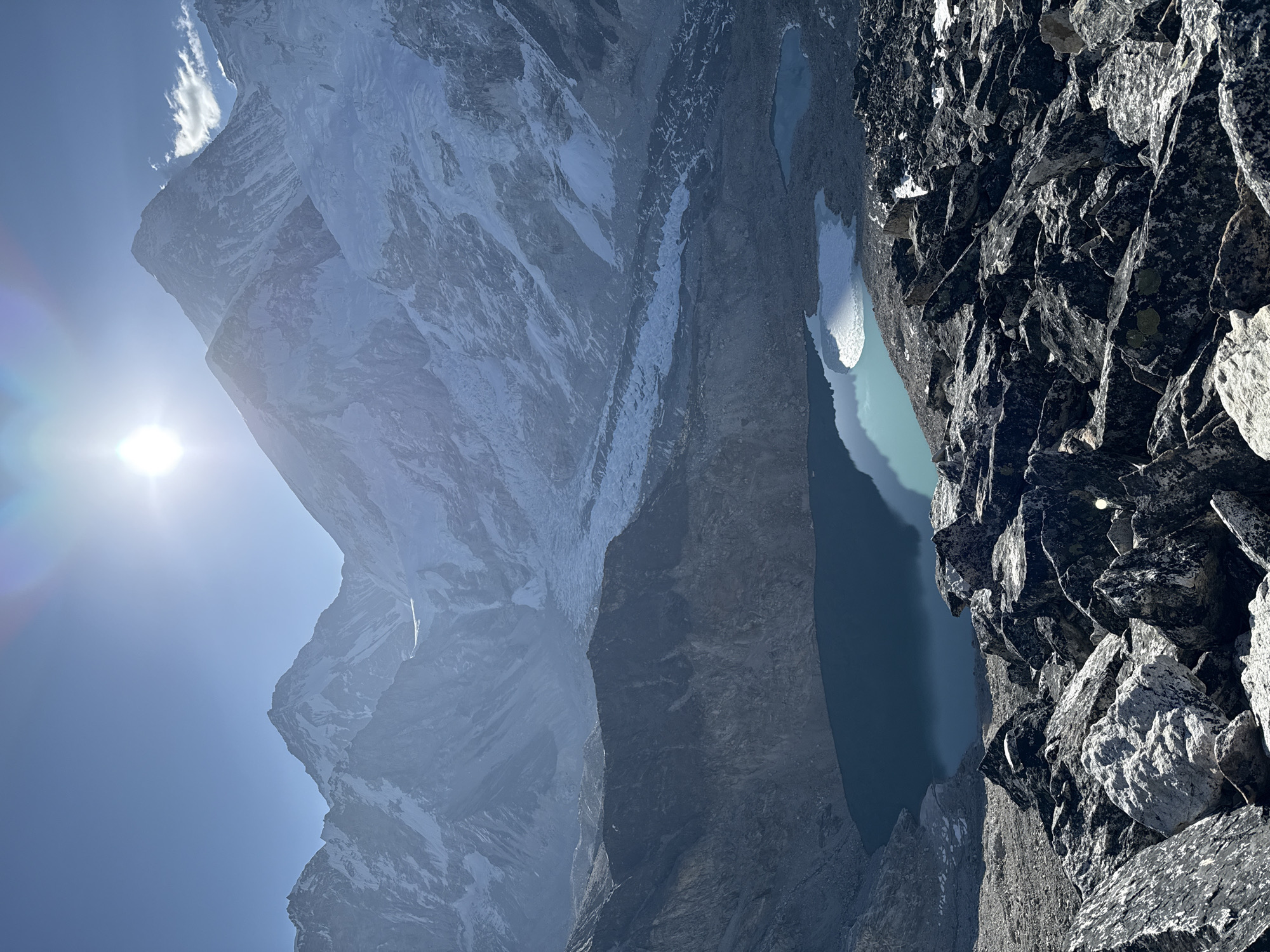This media file captures a majestic, high-altitude mountain landscape, looking down from a steep, rocky vantage point. The scene is dominated by massive, snow-capped peaks that rise dramatically on the left and center, their rugged faces showing both white snowfields and exposed dark rock. These mountains recede in layers, conveying immense scale and depth.
To the right and below the towering peaks, several serene glacial lakes or tarns are visible. Their waters exhibit a striking turquoise color, reflecting the clear blue sky. One of the lakes appears partially covered by ice or snow along its edge.
The immediate foreground on the right is composed of a dark, sharply textured, and steeply angled rocky slope, suggesting the photographer is positioned on a high, craggy ridge or cliff edge overlooking the valley and lakes.
The sky, visible in the upper left, is a vibrant clear blue. A bright, dazzling sun is prominently featured in the upper-left quadrant, casting a strong glare and producing a distinct lens flare, indicating intense daylight, likely mid-morning or early afternoon. A few wispy white clouds are scattered near the mountain summits.
The overall impression is one of immense natural grandeur, remoteness, and pristine beauty, characteristic of an alpine environment like the Himalayas, as suggested by the Khumjung, Nepal context. There is no visible human activity or text in the image.