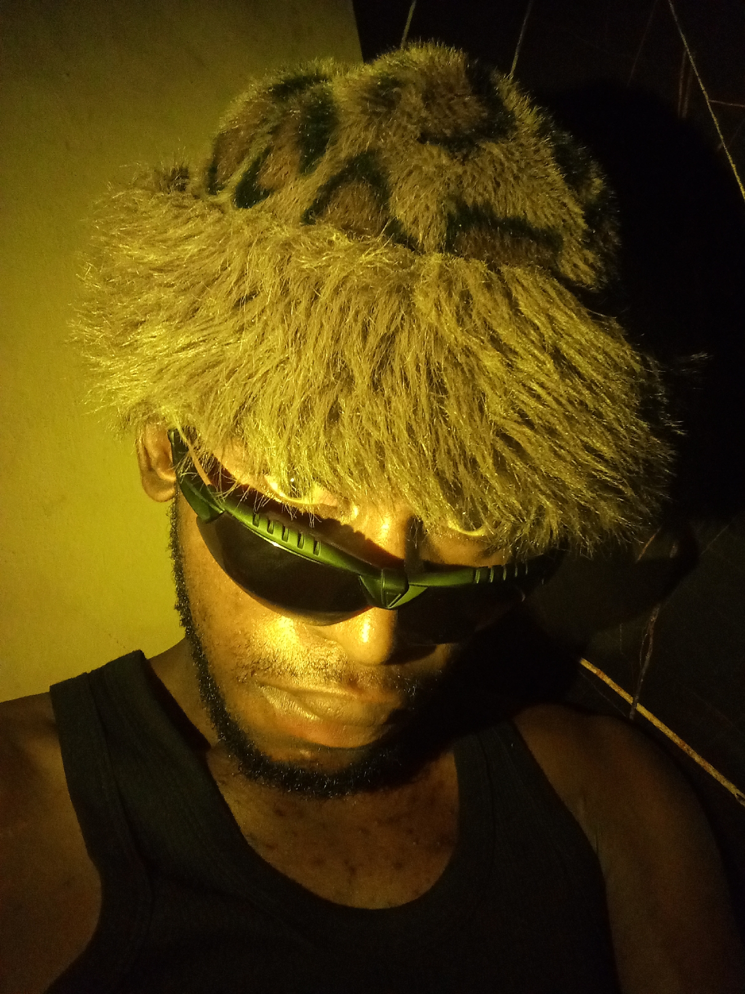 A person, likely male, is shown from the chest up, wearing a fuzzy bucket hat with a leopard print pattern. The hat has a prominent rim of long, light brown fur. The individual is also wearing dark-rimmed sunglasses with green accents. The lighting in the image is a strong yellow hue, casting shadows and highlighting the textures of the hat and skin. The person appears to have a light beard and is wearing a dark tank top. The background is dark and indistinct, with some faint lines suggesting a structure or objects in the environment. The scene is indoors, possibly in a room or a dimly lit space. There is no clear indication of the location within the city of Lokoja, Nigeria.