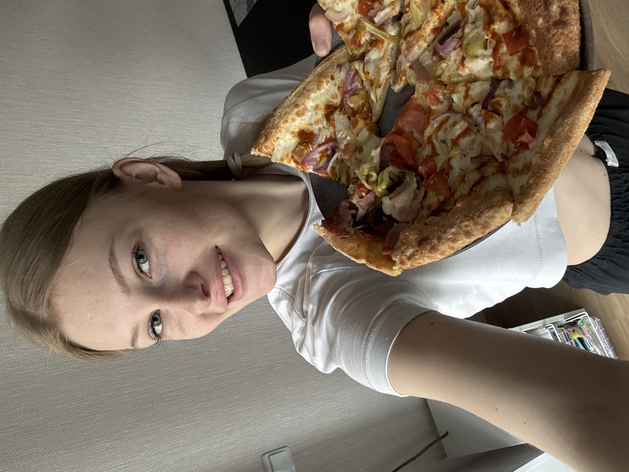 A young Caucasian woman with blonde hair and blue eyes, wearing a white t-shirt, smiles at the camera while holding a pizza. The pizza is on a dark plate and has various toppings, including red onions, tomatoes, and what appear to be peppers. The woman's right arm is visible, and she is wearing a black garment on her lower body. The background consists of a textured grey wall and what looks like a shelf unit on the left side of the image, with some items on it. The lighting suggests an indoor setting, possibly a home. There is no visible indication that this image was taken in the city of Perm, Russia.