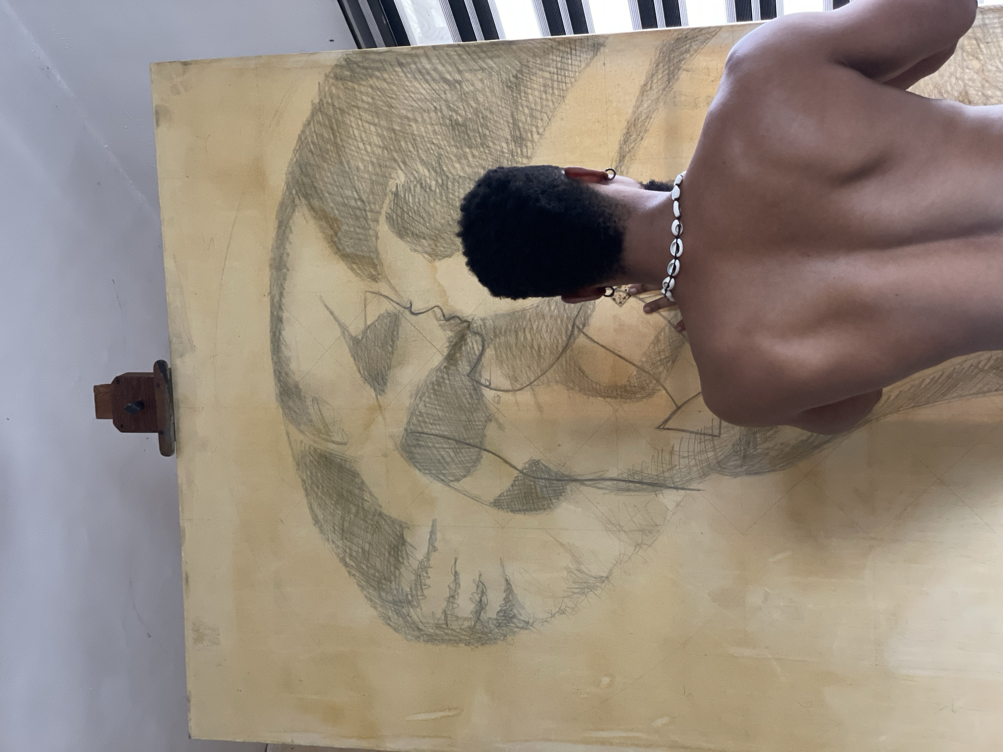An overhead shot captures a person from behind, actively engaged in drawing on a large, light-colored canvas or board. The individual, appearing to be male, has close-cropped dark hair and is shirtless, revealing their back and shoulders. They wear a prominent necklace made of white cowrie shells and a small, gold-colored hoop earring in their left ear.
The large canvas is filled with a detailed pencil sketch, predominantly in shades of gray. The drawing appears to be a human skull or face, intricately rendered with cross-hatching and shading, suggesting a work-in-progress or an expressive artistic style. Construction lines or a grid are subtly visible underneath the main sketch. The board itself shows signs of wear and slight discoloration, consistent with an artist's workspace.
The artwork is propped up, likely on an easel or stand, a small wooden clamp mechanism visible on the left edge of the board. In the background, a plain light-colored wall is visible on the left, and at the top, striped window blinds or curtains indicate an indoor setting during daylight hours. The overall scene suggests a dedicated artist's studio or creative space. There is no visible text in the image.