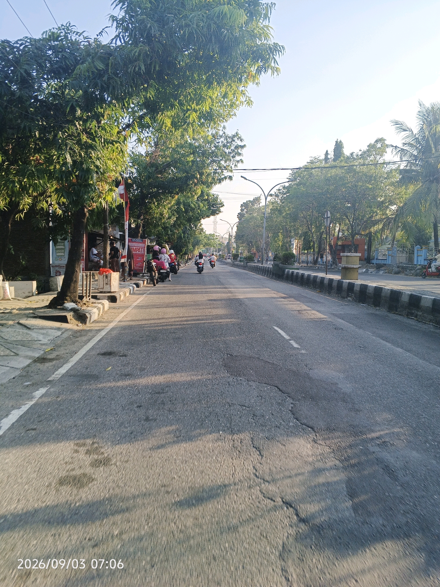 The image depicts a street scene in Pare-Pare, Indonesia, on September 3, 2026, at 7:06 AM. The perspective is from a moving vehicle, likely a motorcycle, looking down a paved road lined with trees on both sides. The sun is low in the sky, casting long shadows across the road and sidewalk.
On the left side of the road, there is a sidewalk with several people gathered near a small kiosk or shop. A few motorcycles are parked along the curb. One motorcycle has a person in a pink helmet and a passenger, while another has two people wearing pink helmets. A red and white flag is visible behind the kiosk.
The road itself is asphalt, with visible cracks and some dark patches, possibly from oil or water stains. There are white painted lines on the road indicating lanes. A concrete barrier with alternating black and white stripes separates the road from the sidewalk on the right side. More trees and some buildings are visible across the barrier on the right. Further down the road, several motorcycles are in motion, heading away from the camera. The sky is clear and bright blue.