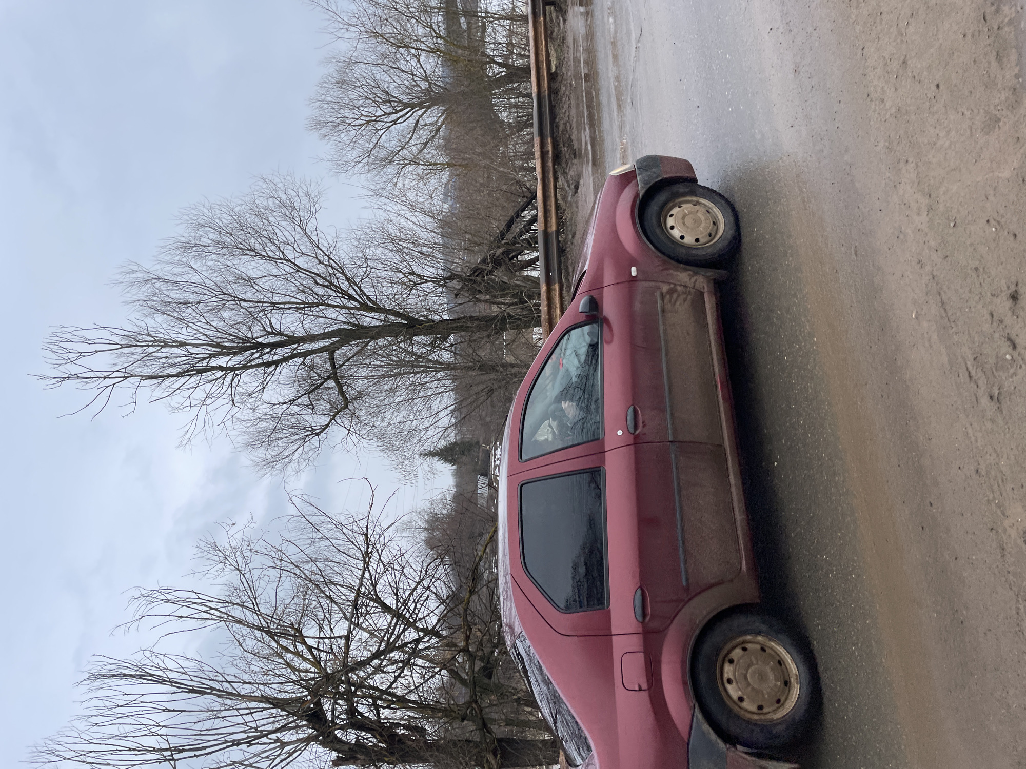 A red car is parked on a muddy road in Фоки, Russia. The day is overcast, with bare trees lining the road. The car is dirty.