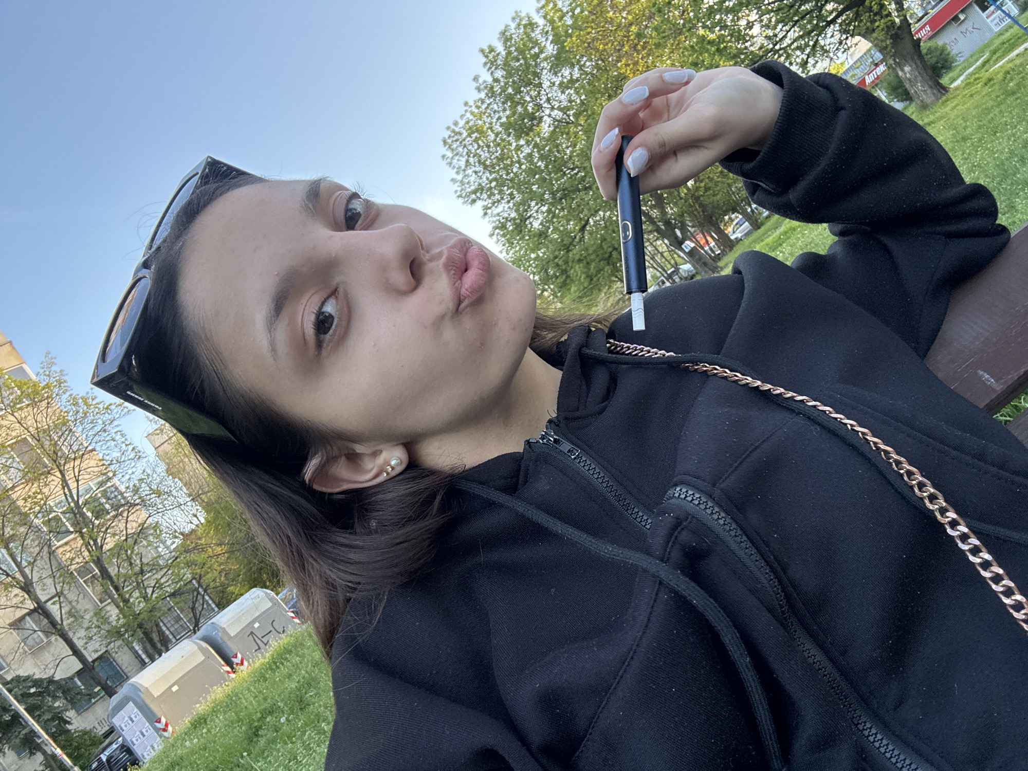 The media file is a close-up selfie of a young woman reclining on a wooden bench outdoors under a bright blue sky. She is seen from a low angle, looking towards the camera with her lips slightly pursed in a pouting expression. She has dark hair, with black sunglasses pushed up onto her head.
The woman is wearing a black zippered hoodie, and a gold-colored chain is visible across her chest. Her fingernails are painted a light, pale color. In her right hand, she holds a dark, cylindrical device, likely a vaping device or e-cigarette, which has a small white tip and a yellow smiley face logo on its side.
The setting appears to be a park or an urban green space. Behind her, there are green trees and multi-story buildings. Two large grey containers marked with "А-С" and other symbols are visible on the left side of the frame. On one of the buildings in the distance, a red sign with Cyrillic text is partially visible, showing "АПТЕКА" (Pharmacy) and "М.К.", which provides a location cue suggesting an urban environment in Sofia, Bulgaria. The image was taken during clear daylight conditions. The overall mood is casual and possibly playful.