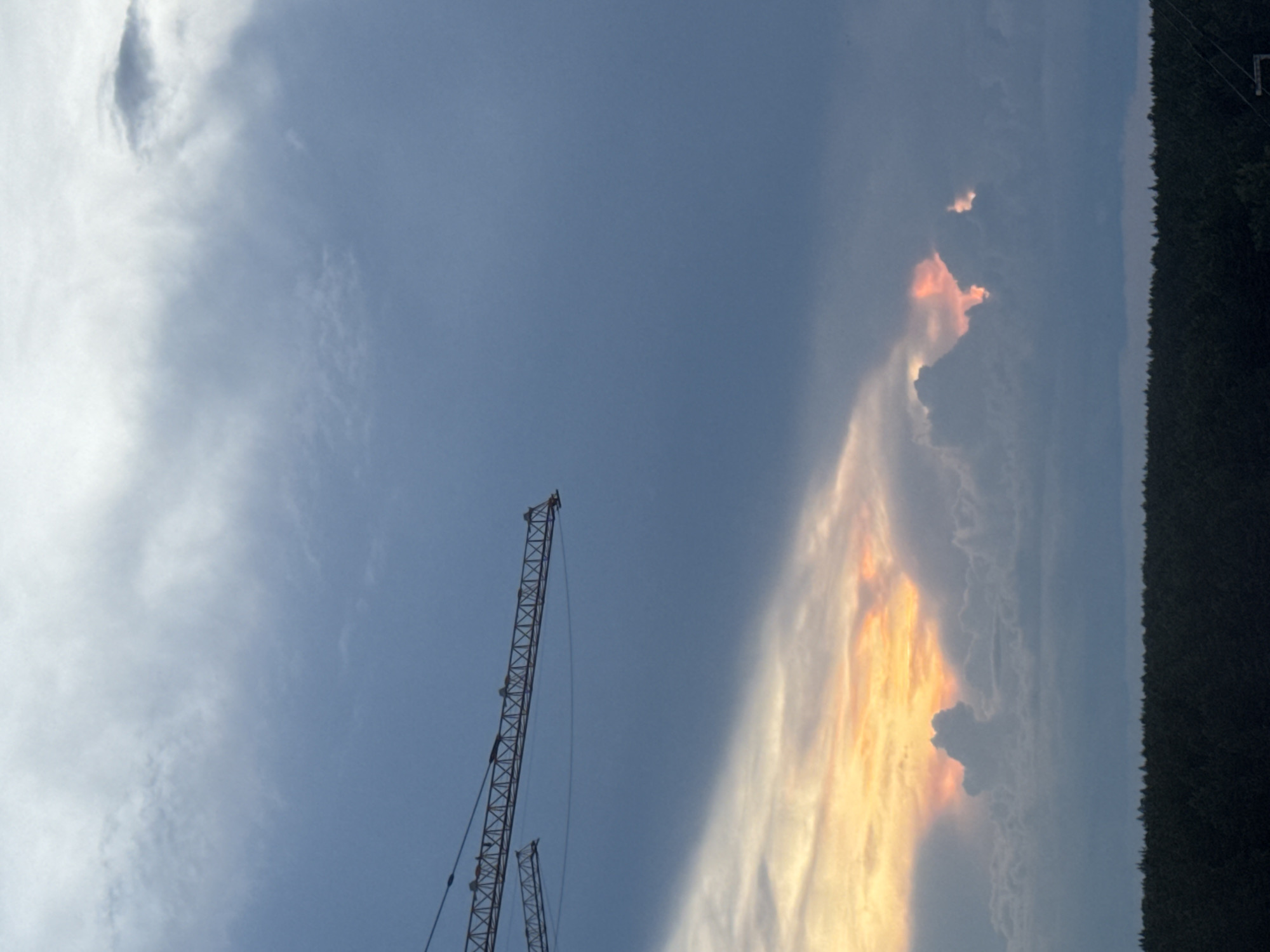 The image depicts a dramatic cloudy sky with the setting sun visible through breaks in the clouds, casting a warm orange and pink glow. In the upper left portion of the frame, the lattice structure of a construction crane is visible against the darker blue sky. Below the colorful sunset clouds, a dense, dark line of trees forms the horizon, likely representing a forest or a treeline on the outskirts of a city. The overall mood is one of impending weather or the transition from day to night. The location is identified as Izhevsk, Russia.