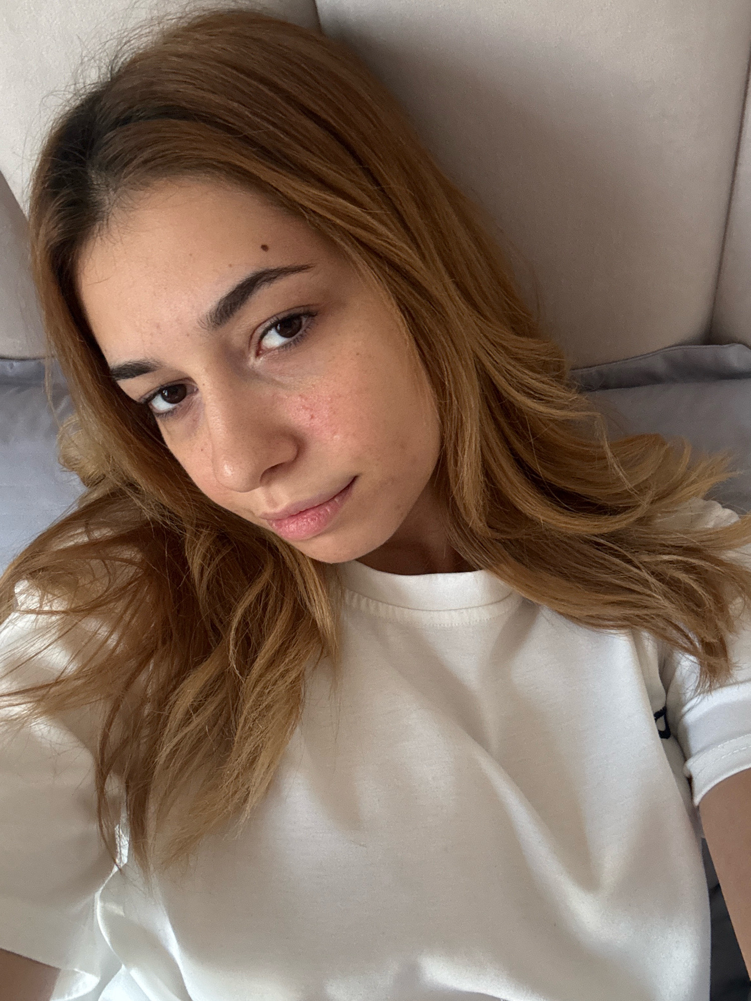 A young woman with light brown, wavy hair and freckled skin is lying down, looking up at the camera. She is wearing a white t-shirt. Her hair frames her face and falls over her shoulders. She has a mole above her right eyebrow and some redness on her cheeks. The background appears to be a bed with a gray pillow. The image was taken in Getahovit, Armenia.