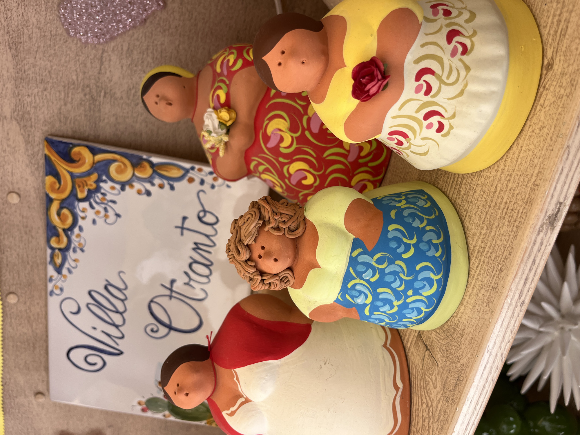 The image shows four small, hand-painted ceramic figures of women, seemingly arranged on a shelf along with other decorative items. The figures are rotund and stylized, with smooth, featureless faces except for small indentations indicating facial features. They are each painted with different colored dresses and patterns. Next to the figures is a ceramic tile or plaque with the inscription "Villa Otranto" in cursive script, framed by a blue and yellow decorative border. Otranto is a town in the Apulia region of Southern Italy. Other items are slightly visible in the background, appearing to be decorative plants, and a sparkly object. The scene is brightly lit and seems to be a display of items for sale, or part of a domestic decoration.