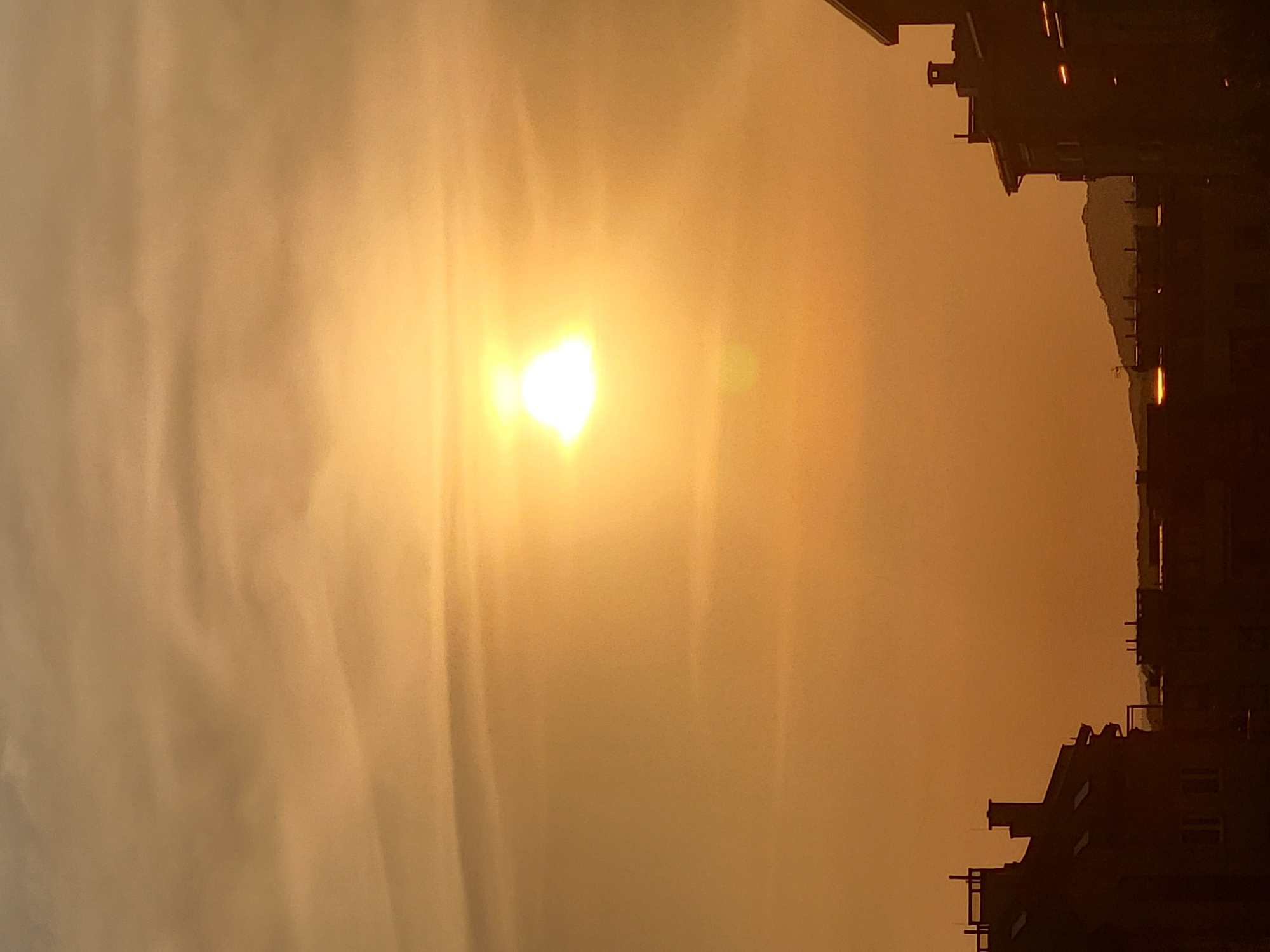 The image depicts a sky filled with a warm, golden sunset glow, with the sun prominently visible as a bright, luminous orb. Wispy clouds are scattered across the sky, adding texture and depth to the scene. In the foreground, the silhouetted rooftops of buildings are visible, suggesting an urban environment. Some of these buildings feature chimneys and architectural details that are rendered in dark tones due to the backlighting from the sunset. In the distance, a gently sloping hillside is discernible against the horizon. The overall impression is one of a serene evening in a city, bathed in the warm light of the setting sun.