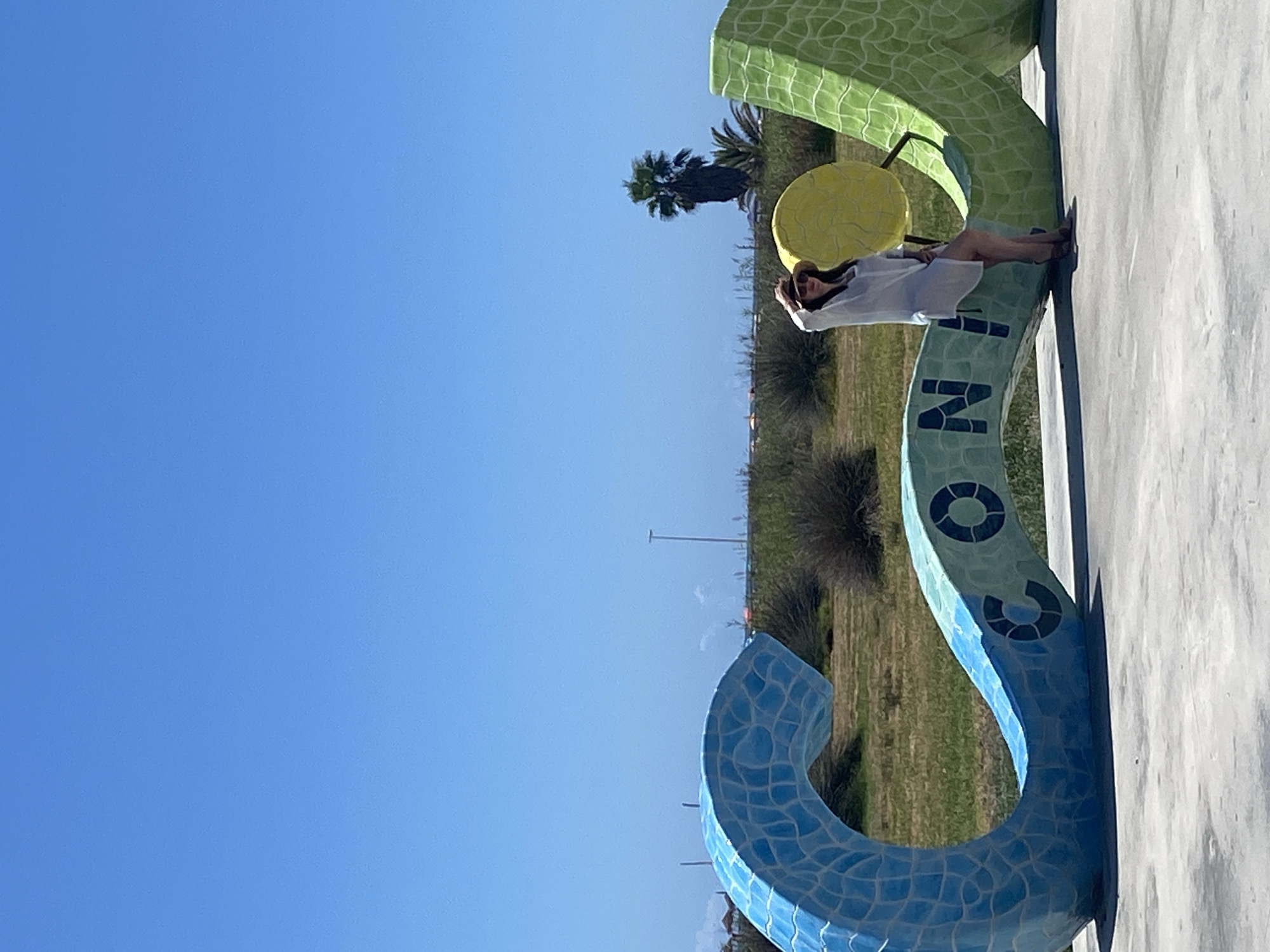 A woman wearing a white cover-up and a straw hat poses for a photo in front of a large, colorful, mosaic sculpture that spells out "CONIL". The sculpture is a vibrant blue on the left, transitioning to light blue and green, with the letters "CONIL" visible in dark blue. To the right of the letters, there's a yellow circular element and a green serpentine shape. The woman is leaning against the sculpture, her hand on her hat, looking towards the camera. The background consists of dry vegetation, a palm tree, and a clear blue sky. In the distance, there are hints of buildings and infrastructure, possibly a road or coastline, suggesting an outdoor, possibly coastal, setting. The ground in the foreground is a paved surface. The overall scene conveys a bright, sunny day in a public outdoor space, likely a promenade or park in Conil De La Frontera, Spain, given the sculpture's lettering.
