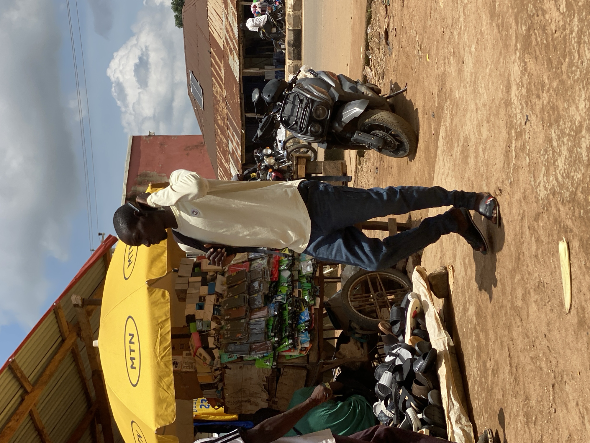 The image depicts a street scene in Kafanchan, Nigeria, under a bright, cloudy sky. In the foreground, a young man with dark skin is talking on a mobile phone, holding it to his right ear with his right hand. He is wearing a light-colored long-sleeved shirt and dark blue jeans, with black sandals on his feet. He carries a dark backpack over his left shoulder.
To his left, a market stall is set up under a large yellow umbrella with the "MTN" logo. The stall is filled with an assortment of goods, primarily mobile phone accessories such as cases and screen protectors, neatly displayed. Beneath the stall, a pile of black and white sandals is arranged on a light-colored cloth. Another person, partially visible on the far left, wears a dark shirt and is looking towards the market stall.
Further into the background, a parked scooter with a dark grey body and black accents is prominently displayed. Behind the scooter, a street runs across the image. Several other motorcycles are visible parked along the street, and some people are seen near them. Buildings with corrugated tin roofs, some showing signs of age and wear, line the street. One building has a red facade, and another features solar panels on its roof. The ground is covered with reddish-brown dirt.