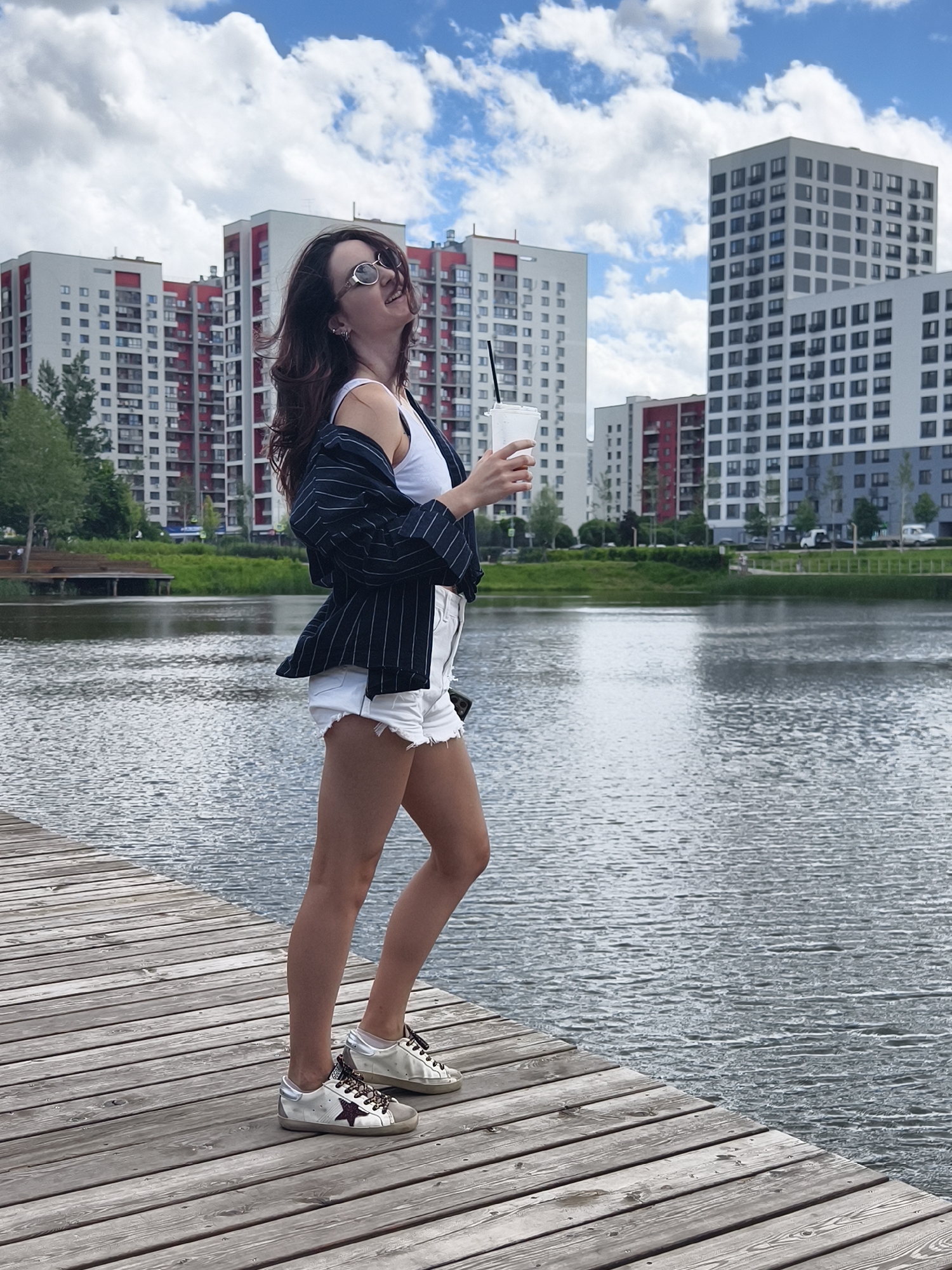 A young woman with dark hair is standing on a wooden pier next to a body of water, holding a cup with a straw. She is wearing sunglasses, a white tank top, a dark pinstripe blazer draped over her shoulders, and white distressed denim shorts. Her sneakers are white with gold and purple star accents. In the background, several modern residential buildings with red and white accents are visible. The sky is blue with white clouds. The scene is outdoors, suggesting a city park or waterfront area. Based on the architecture, the location is identified as the city of Tyumen, Russia.