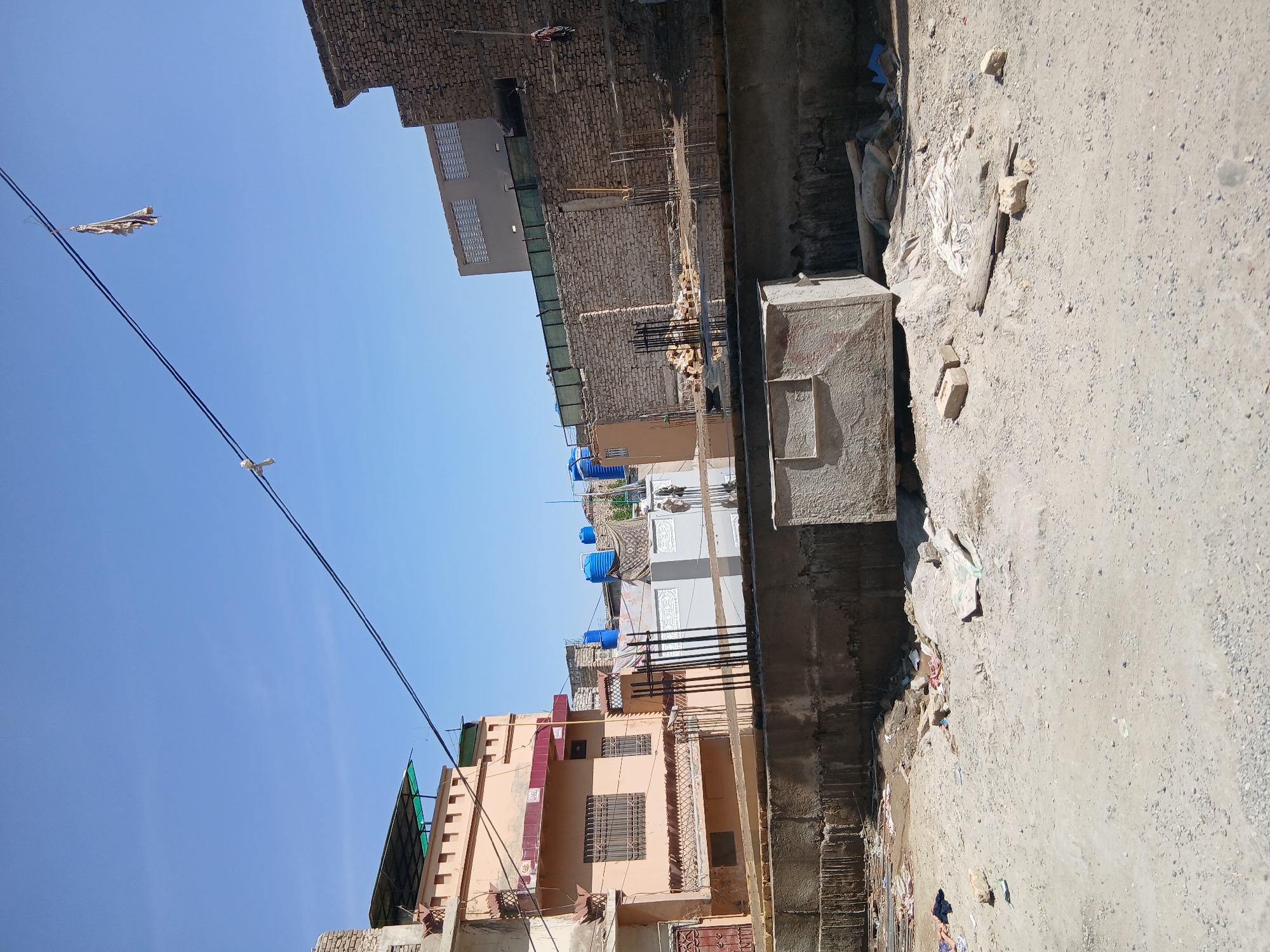 This image captures a street scene in Quetta City Tehsil, Pakistan, under a clear, bright blue sky.
The scene depicts a narrow, unpaved street or alleyway, with buildings lining both sides. On the left, several multi-story buildings are visible; one is a distinctive peach color with grilled windows and balconies, while another further back is lighter grey with intricate grate-like patterns. Multiple blue plastic water tanks are perched on the rooftops of these structures. Some buildings show signs of ongoing construction or partial disrepair, with exposed brickwork and rebar visible.
Overhead, power lines stretch across the sky, and a piece of tattered fabric, possibly a torn kite, is snagged on one of them. The ground on the right side of the image is an unpaved, dusty surface composed of dirt and gravel, littered with construction debris, loose bricks, and some trash. A large, weathered concrete block rests on this uneven ground.
The overall impression is that of an urban or semi-urban environment characterized by dense housing and ongoing development. The bright, cloudless sky indicates it is daytime, likely mid-morning or afternoon. No people are visible, nor is any legible text apparent on buildings or signs.