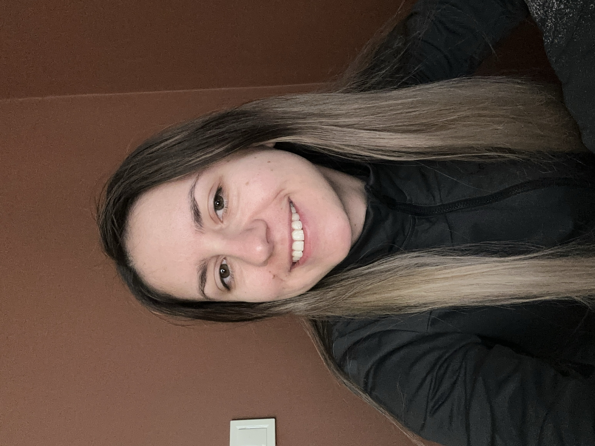 A young woman with long brown and blonde hair is smiling at the camera. She is wearing a black zip-up jacket. The background is a brown wall with a white electrical outlet visible at the bottom. The location is not discernible from the image.