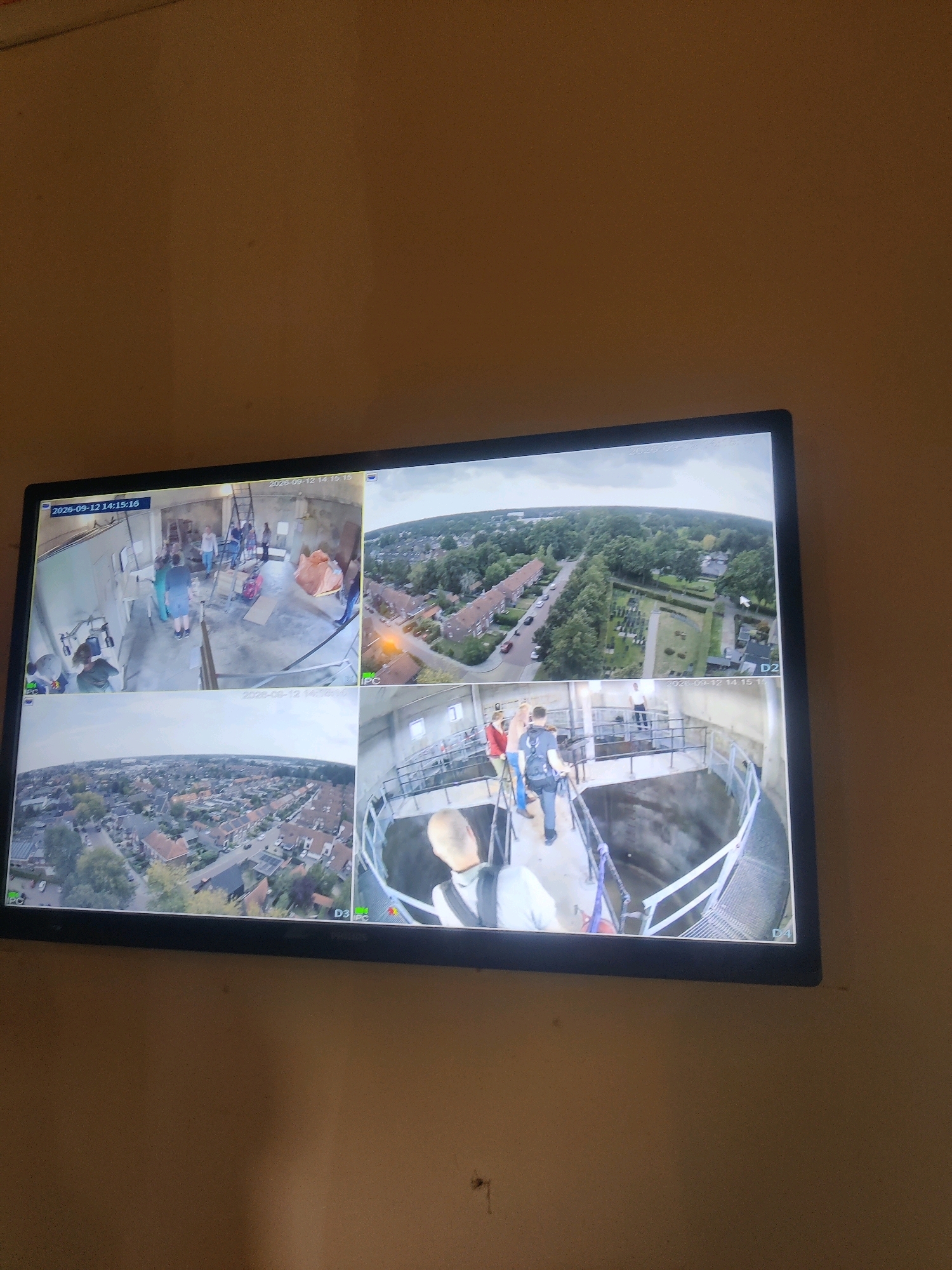 The image displays a four-quadrant CCTV monitor showing various scenes.
The top-left quadrant shows an indoor scene with several people, some appearing to be workers, in a room with concrete walls and floors. There are construction materials and tools visible.
The top-right quadrant shows an aerial view of a suburban area with houses, trees, and a road. A bright orange object is visible on the road.
The bottom-left quadrant also presents an aerial view of a residential area, similar to the top-right quadrant, with houses, gardens, and trees.
The bottom-right quadrant shows an outdoor scene with a group of people on a metal walkway or platform surrounding a large, deep opening. Some individuals are wearing harnesses, suggesting a controlled activity like rappelling or bungee jumping. The environment appears to be a structure with industrial elements and a concrete interior.
The timestamp "2023-09-12 14:15:16" is visible in the top-left and top-right quadrants. "2023-09-12 14:15:16" is also visible in the bottom-left quadrant, and "2023-09-12 14:15:15" in the bottom-right quadrant. Camera labels "D2", "D3", and "D4" are visible in the bottom-right and bottom-left quadrants. "IPC" is visible in the top-right and bottom-left quadrants.
Based on the context provided by previous queries (which are not visible here but implied by the instruction to include location details), this footage likely originates from Winterswijk, Netherlands. The scene in the bottom-right quadrant, showing people on a platform overlooking a deep shaft, could potentially be related to a jump facility or a similar recreational or industrial site in or around Winterswijk. The aerial views would then correspond to the surrounding urban and suburban landscape of the area.