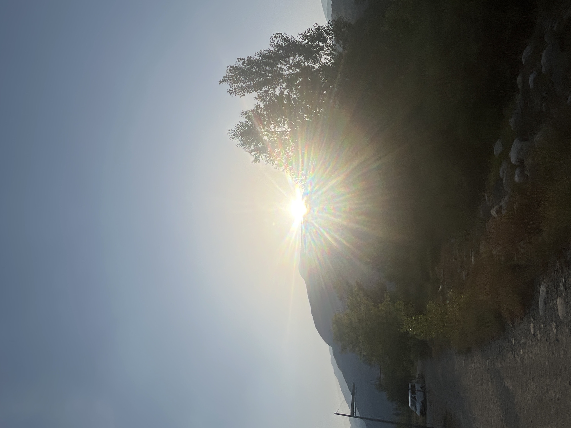 The image captures a sunburst effect behind silhouetted trees and mountains, possibly in the vicinity of Benasque, Spain. A gravel path curves into the foreground on the left, where a white van is parked. The sky is a pale blue, fading to white near the horizon, with the sun's rays creating a rainbow effect as it peeks over a mountain range. The surrounding landscape is dominated by dense, dark foliage and rocky terrain.