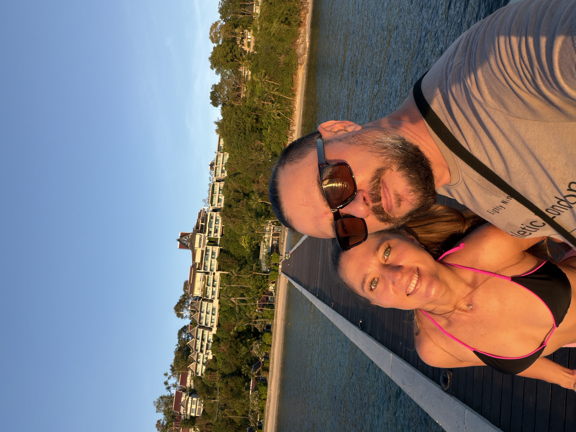 In Baan Taling Ngam, Thailand, a couple stands on a wooden pier extending over calm, blue water. The woman, on the left, wears a black bikini with pink trim and smiles at the camera. She has long, brown hair and green eyes. The man, on the right, sports sunglasses and a beard. He is wearing a light-colored t-shirt with text that is partially visible. The background features a lush, green hillside covered with trees and buildings, indicative of a resort or hotel. The sky above is a clear, bright blue, and the sun, likely setting, casts a warm glow on the scene, creating shadows on the pier and highlighting the couple's faces. The overall atmosphere is one of leisure and vacation.