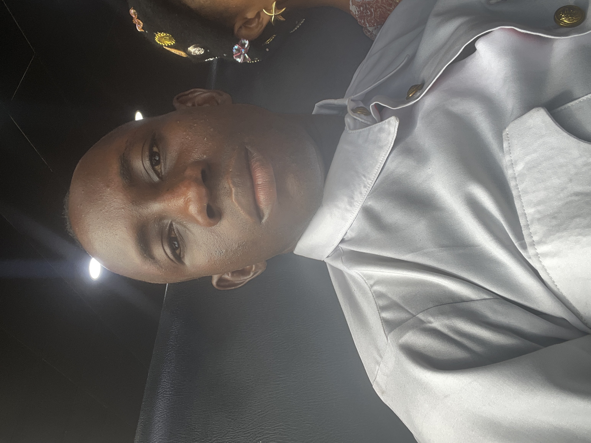 A close-up shot shows a dark-skinned person wearing a white collared shirt with gold buttons. A portion of another person’s head with decorative hair accessories is visible in the background. The environment appears to be indoors. There is insufficient information to confidently determine the location in Lagos, Nigeria.