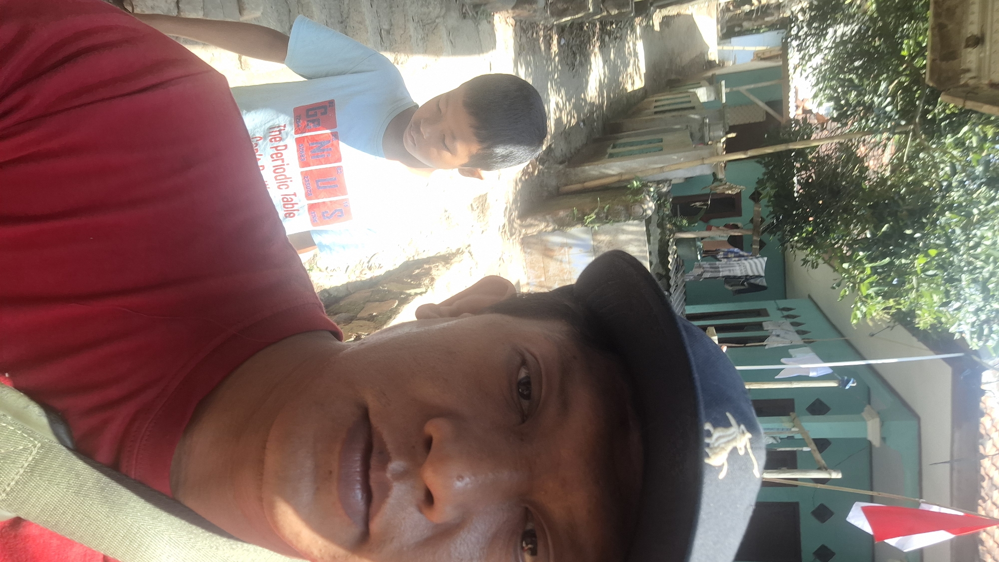 In Cilaku, Indonesia, an outdoor selfie captures two individuals. The foreground is dominated by the face of an adult male wearing a red t-shirt and a dark baseball cap adorned with a silver emblem of what appears to be a horse. His head is turned towards the camera, with his right ear and shoulder visible. Behind him, a young boy wearing a light blue t-shirt with the words "The Periodic Table" and "GENIUS" in large red letters stands looking downwards. The boy's t-shirt displays chemical element symbols and atomic numbers. The background reveals a residential street lined with houses and a fence. One house is painted a light turquoise color, with laundry hanging on a clothesline and an Indonesian flag visible on a flagpole. Sunlight casts shadows on the ground, suggesting a bright day. The ground is a mixture of dirt and stone.