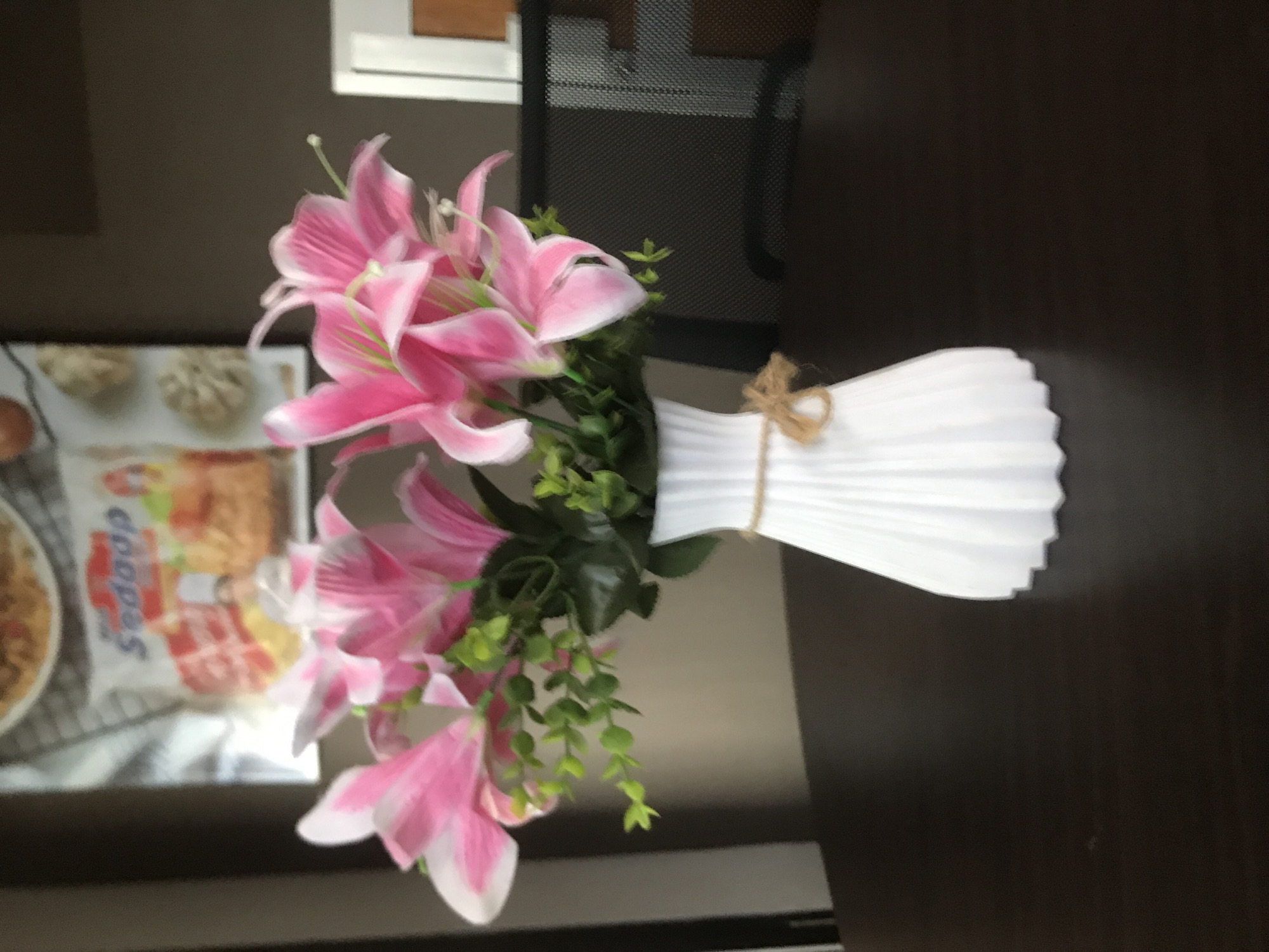 A top-down view of a pink lily arrangement in a white, pleated vase on a dark surface. A package labeled "Sedap" and several food items are visible in the background, and a metal chair is also partly visible. The location is indeterminate and cannot be specified as Lhokseumawe, Indonesia based on this image.