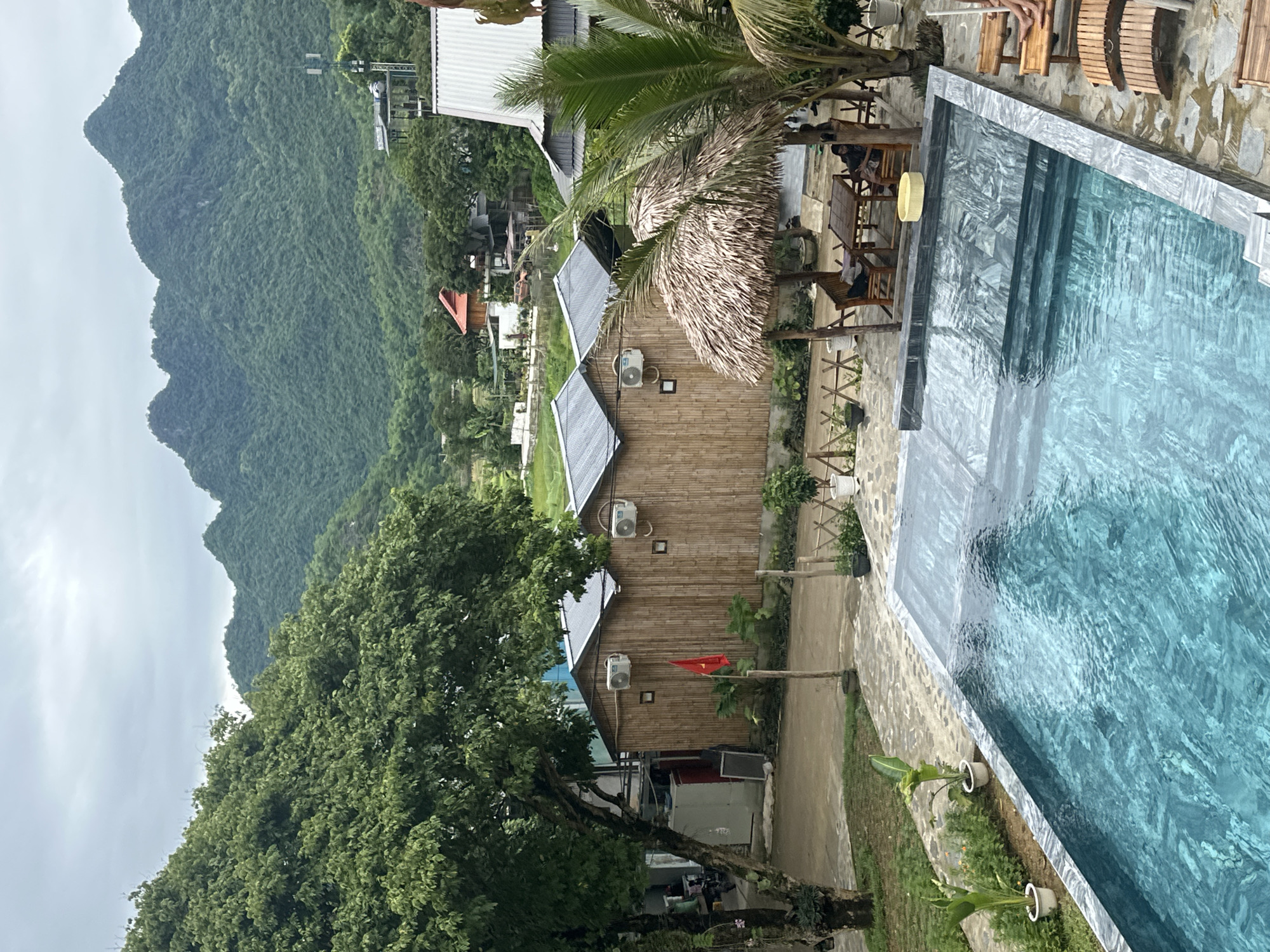 The image depicts a resort or guesthouse in Xóm Khán, Vietnam, set against a backdrop of lush, green mountains. In the foreground, a clear blue swimming pool with stone tiling occupies the lower portion of the frame. To the right of the pool are several wooden lounge chairs, with a person partially visible sitting in one. Adjacent to the pool is a thatched-roof seating area with wooden tables and chairs, where two individuals are seated. Behind this area, several bamboo-sided structures with metal roofs, likely guest rooms or bungalows, are visible. Each structure has an air conditioning unit mounted on the exterior wall. A Vietnamese flag is planted in front of one of these buildings. A large, leafy tree stands to the left of the structures, and beyond that, a rural landscape with scattered buildings and more greenery stretches towards the mountains. The sky is overcast.