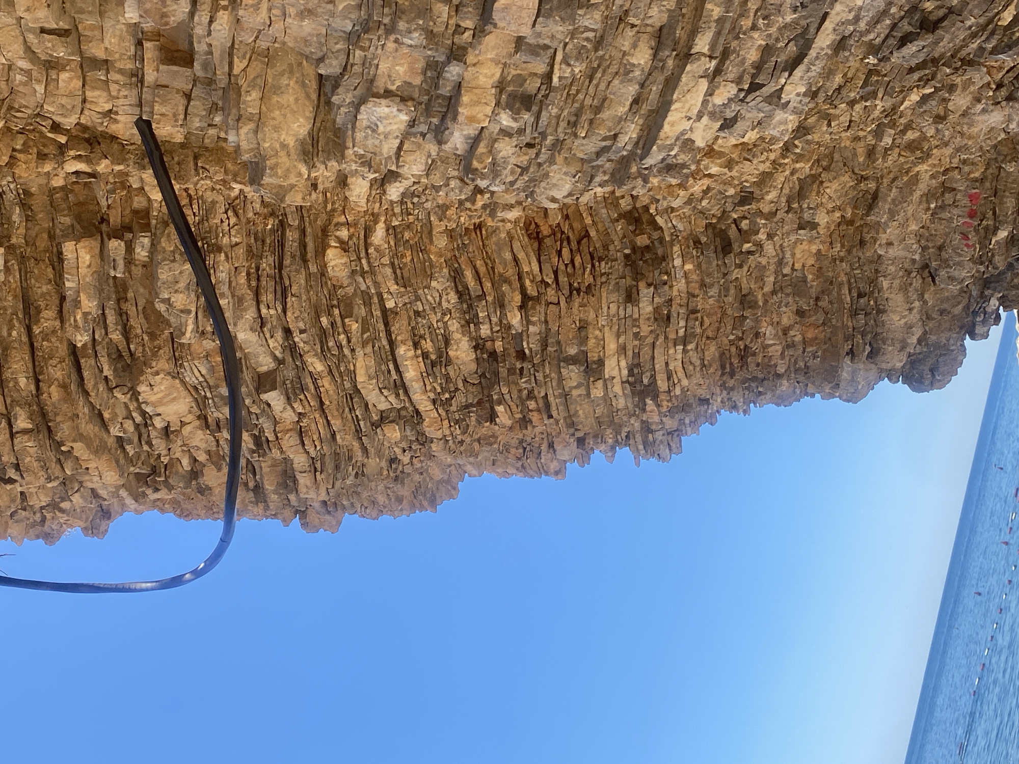 The image shows a rock overhang in Budva, Montenegro. The layered rock formation is tan and brown, with a black cable running along the underside. Below, the sky is a clear, bright blue. To the right, a sliver of water and a row of red and white buoys are visible.