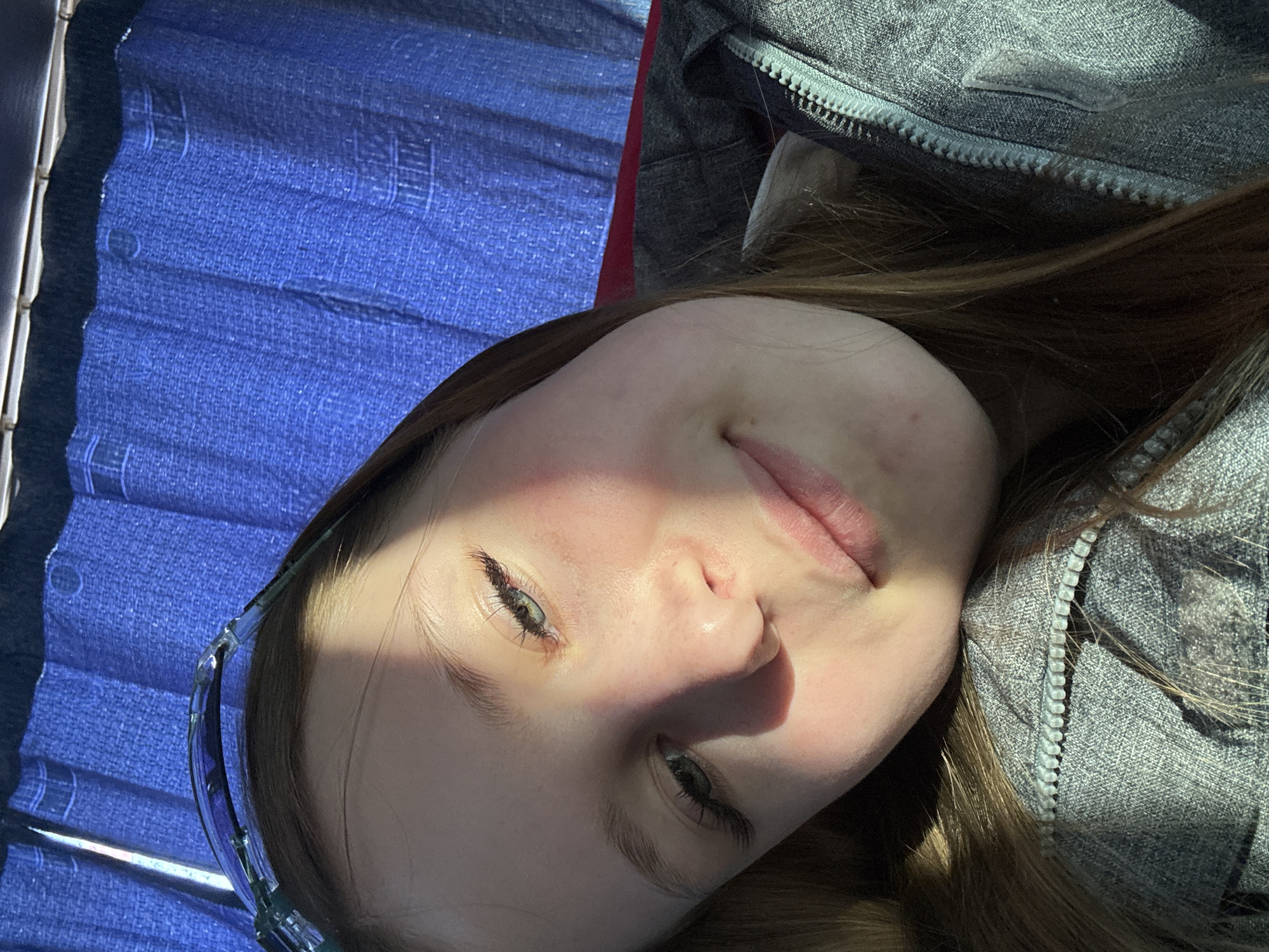 A young woman with fair skin and brown hair is lying down, wearing glasses and a gray zippered jacket. The background includes a blue patterned seat cover, possibly in a vehicle. There is no verifiable information in the image to specify a location within the city of Berezniki, Russia.