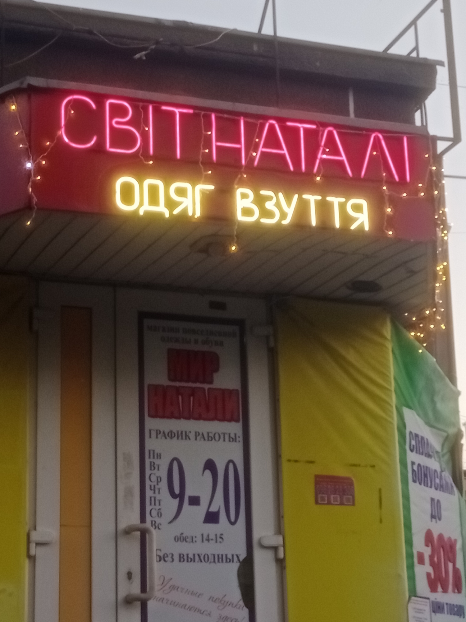 The image shows a storefront in Horishni Plavni, Ukraine. Above the entrance, a neon sign in pink and yellow letters reads "СВІТ НАГАЛІ ОДЯГ ВЗУТТЯ", which translates to "Naked World Clothes Shoes". Below this, a poster displays "МИР НАТАЛИ" ("Natalia's World") with opening hours "9-20" and "обед: 14-15" ("lunch: 14-15"). There is also text that translates to "Without days off". To the right of the entrance, a banner advertises a sale of "-30%". The overall scene depicts a retail establishment selling clothing and footwear.