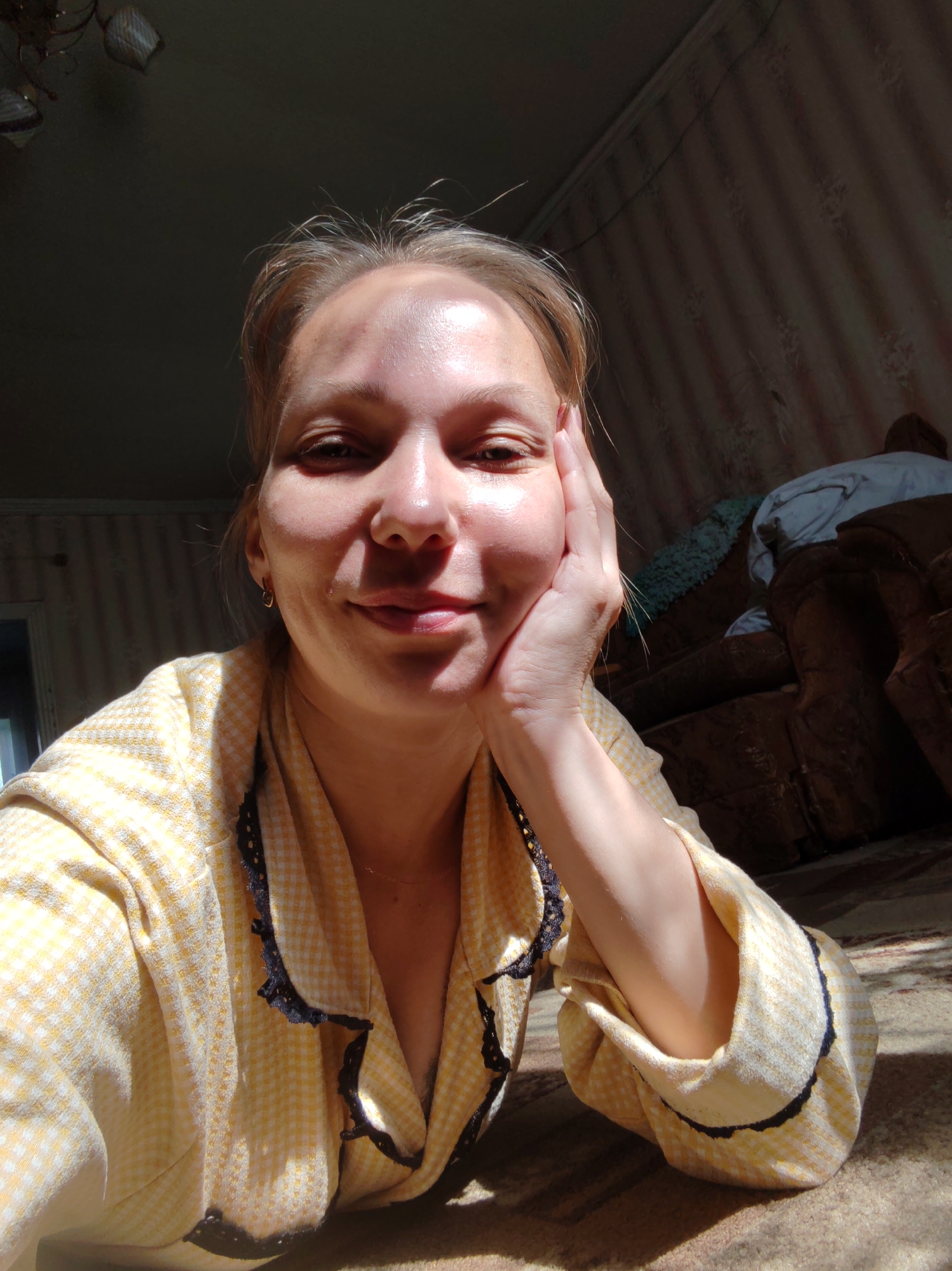 A woman, appearing to be in her late 20s to early 30s, is captured in a close-up, head-and-shoulders selfie taken indoors. She has light skin and blonde hair, which is pulled back from her face. Her left hand is resting on her cheek, and she is looking directly at the camera with a slight smile. She is wearing a yellow and white checkered robe with black lace trim around the collar and cuffs. Sunlight is streaming in from the left, casting a strong shadow across the left side of her face and illuminating the right side.
The background suggests a domestic setting. To the right, a dark-colored sofa with a patterned upholstery is visible, partially covered with blankets or throws. The wall behind the sofa has a striped wallpaper pattern. The floor appears to be carpeted. The lighting and general decor suggest a casual, lived-in environment. The photograph is likely taken in a residential room, potentially a living room or bedroom, within a house or apartment.
The specific location provided, "Chukhlem, Russia," indicates the general geographical area. While the image itself does not contain explicit markers of this location, the details of the interior suggest a home that could be found in a Russian town like Chukhlem. The style of the wallpaper, the furniture, and the overall ambiance are consistent with domestic interiors that might be found in many parts of Russia.