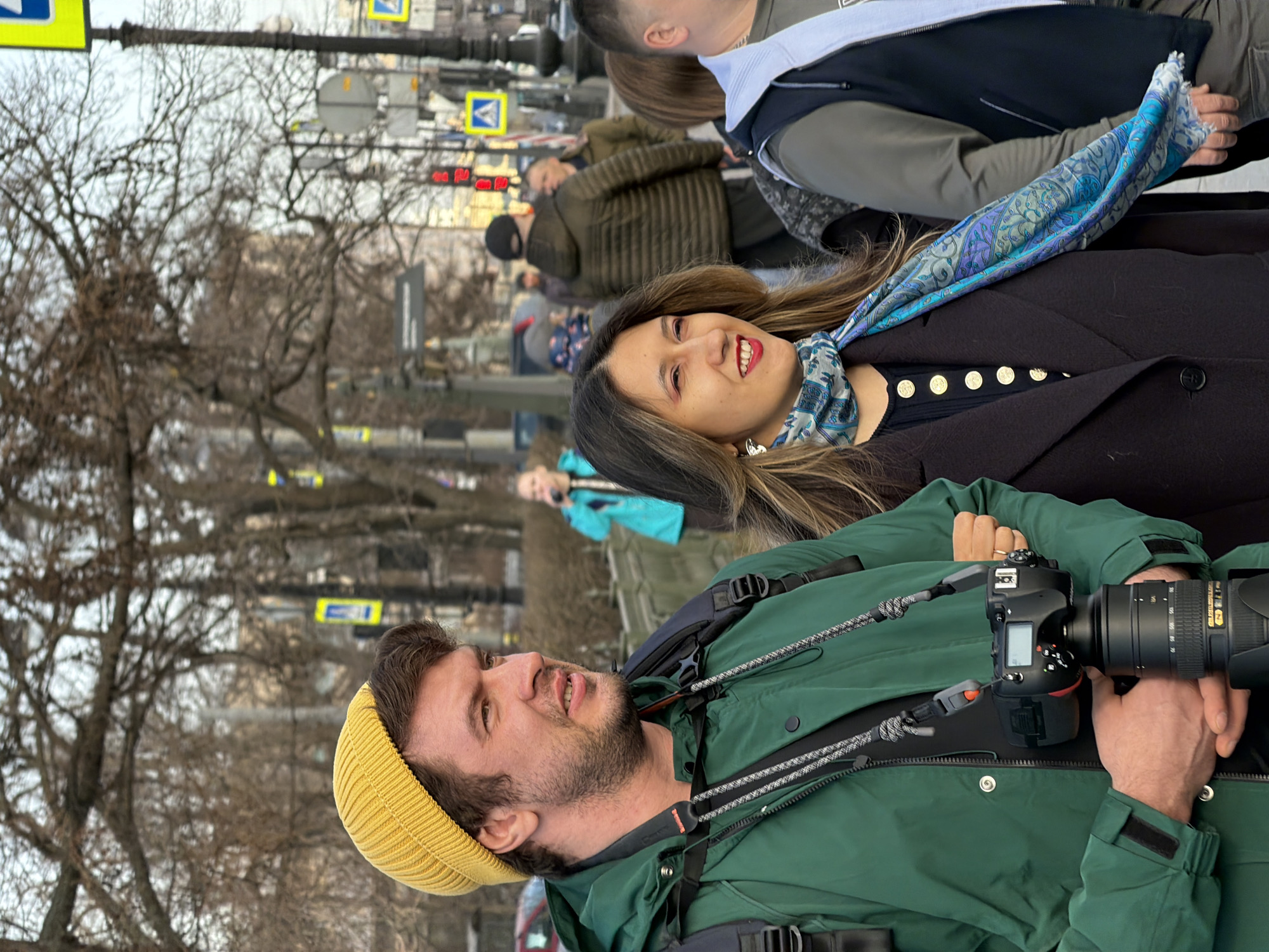 The image depicts a street scene in Saint Petersburg, Russia, characterized by bare trees suggesting a colder season.
In the foreground, a man and a woman walk arm-in-arm. The man, on the left, wears a bright yellow knit beanie, a dark green jacket, and a black backpack. He has short dark hair and light stubble. He holds a professional-grade black DSLR camera with a large lens in his right hand, secured by a braided strap around his neck. His mouth is slightly open, and he is looking towards his right. The woman, on the right, wears a dark coat with gold buttons, a patterned blue and white scarf, and has long, wavy dark hair. She wears red lipstick and a small earring on her visible ear, smiling and looking in the same direction as the man.
In the mid-ground and background, several other pedestrians are visible, blurred. To the left, a woman in a bright blue jacket appears to be holding an object to her mouth. To the right, a man in an olive green puffer jacket and a dark cap walks away from the viewer. The environment includes bare trees, a stone barrier, streetlights, a functional red traffic light, and multiple blue pedestrian crossing signs with white pedestrian symbols on yellow backgrounds, typical of Russian street signage. Faint building structures are discernible in the distant background.