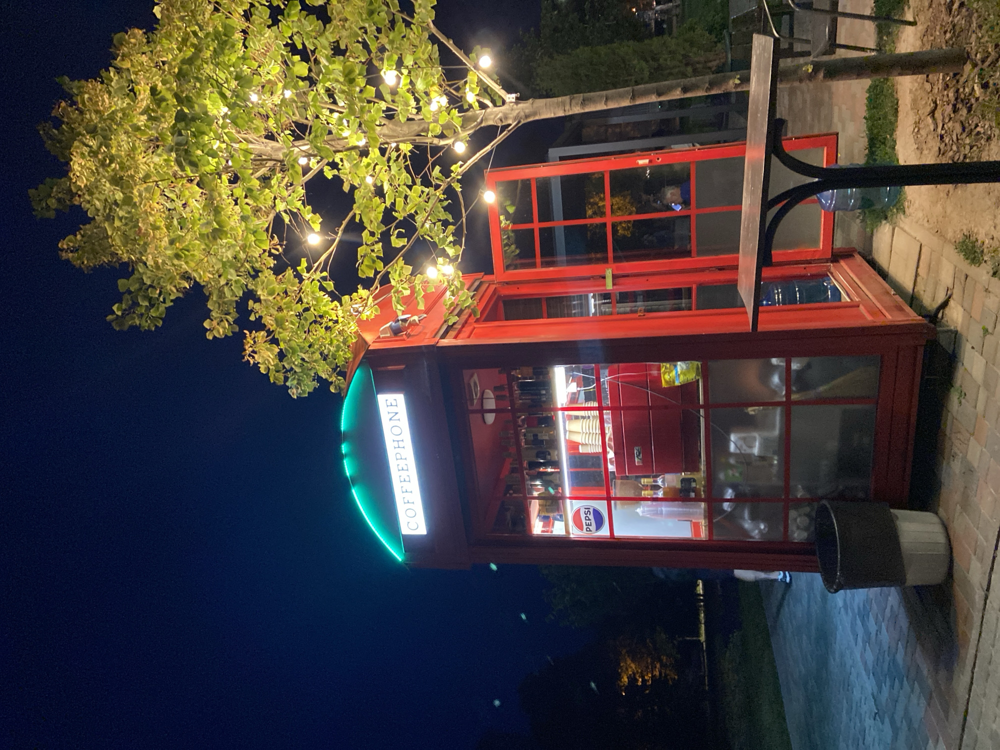 The image shows a red telephone booth converted into a coffee stand, labeled "COFFEEPHONE" in white lettering under a green illuminated arch. The booth is open with its door ajar, revealing a coffee machine, cups, bottles of syrup, and a Pepsi logo visible inside. A tree decorated with string lights stands to the right of the booth, casting a warm glow. In the foreground, a dark-colored trash can sits on a cobblestone path. In the background, a dark blue night sky is visible, along with some trees and a low-lying fence. The scene is likely set in a public outdoor area, possibly a park or a street, in the city of Poltava, Ukraine.