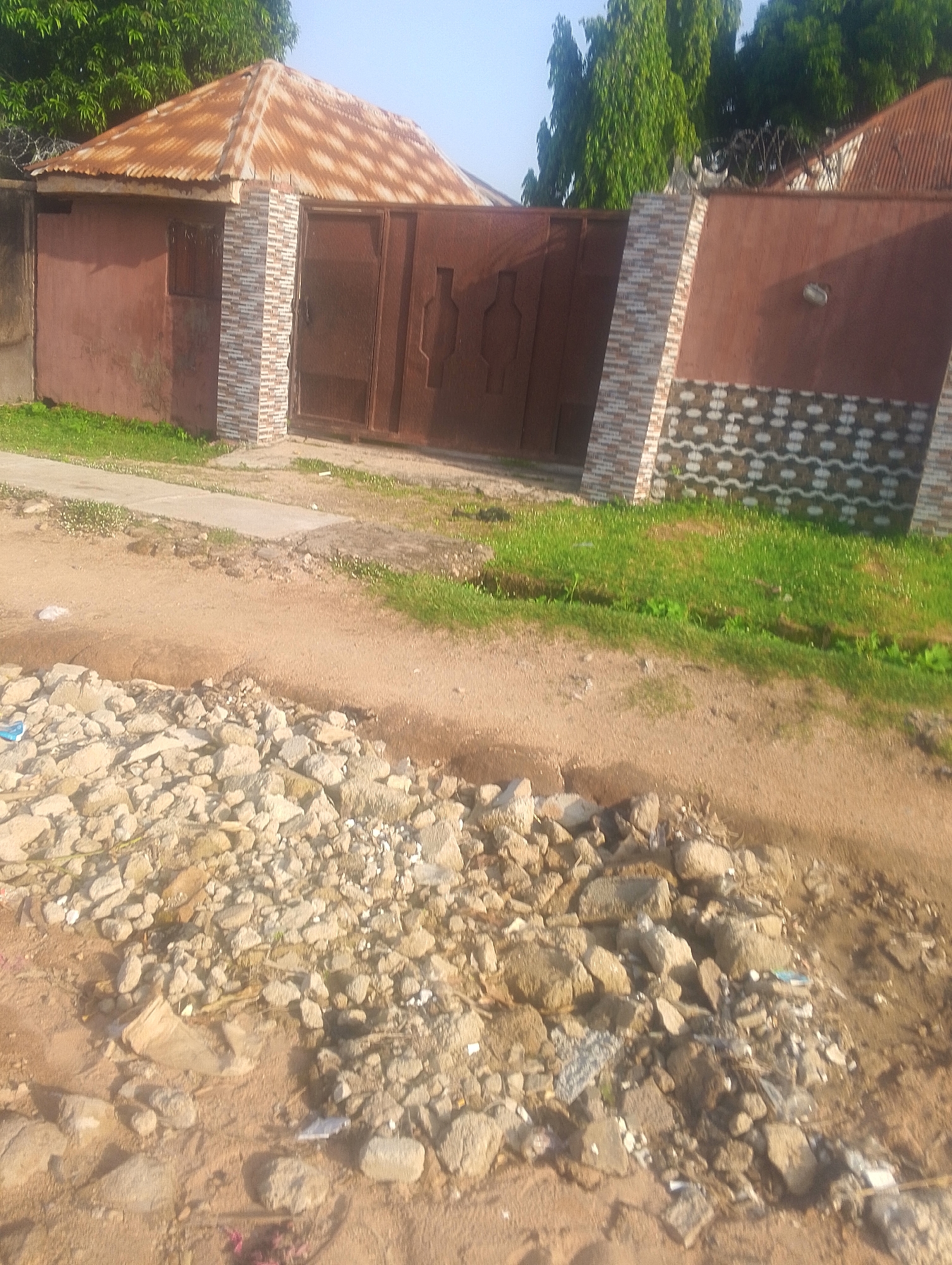 The image shows a front view of a residential property in Jalingo, Nigeria. The property has a large, rusty metal gate and a dark red wall. The gate is flanked by stone-clad pillars. A portion of the wall features a decorative pattern of white and dark grey shapes. Behind the wall, some greenery is visible, including trees and grass. In the foreground, there is a dirt road with a pile of rocks and debris. The overall impression is of a simple, somewhat unkempt residential area.