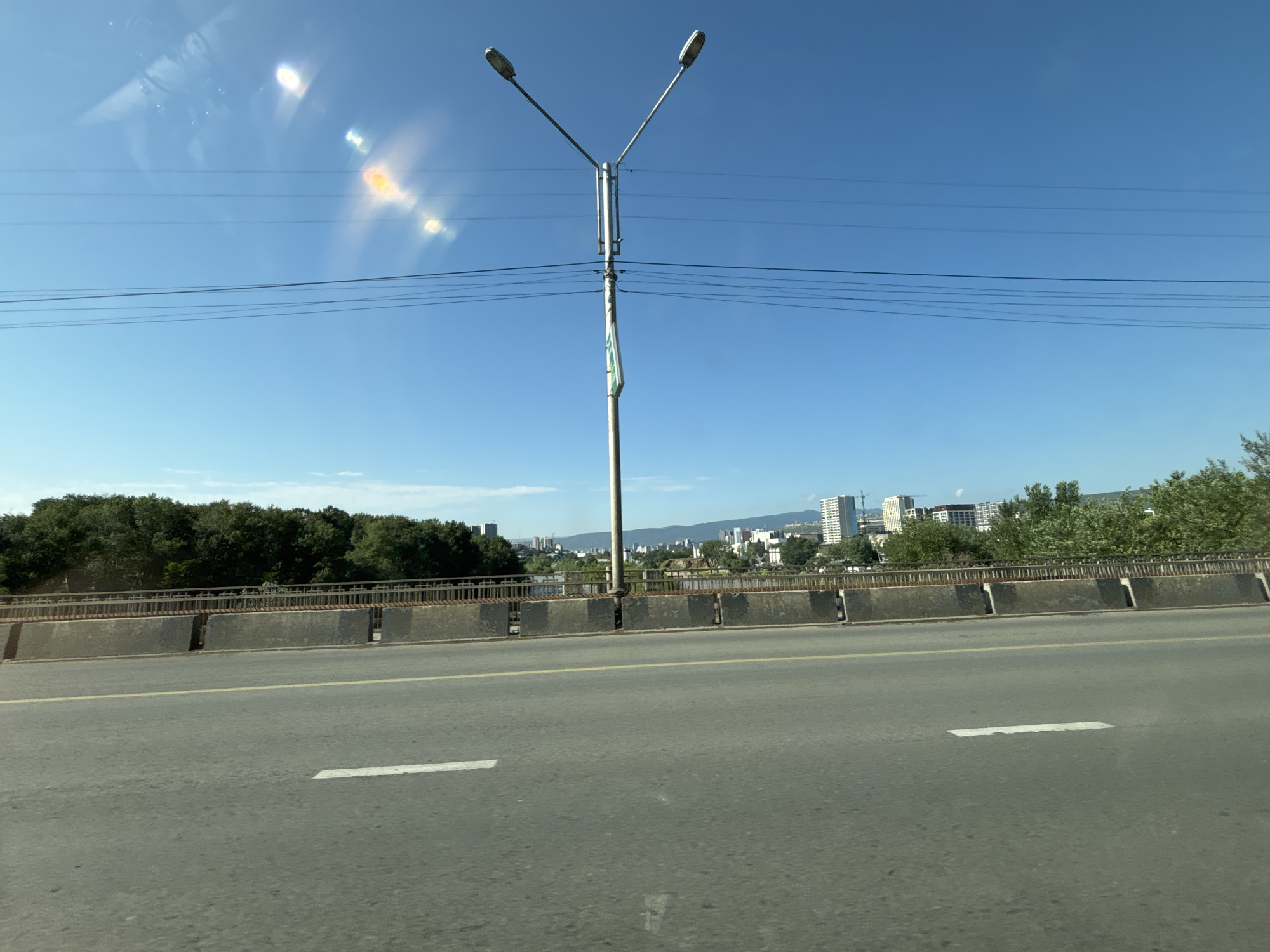 This is a photograph taken from a vehicle moving on a road that appears to be an elevated highway or bridge in Tbilisi, Georgia. The road surface is asphalt with a yellow line marking the center and white lane markings. A concrete barrier with metal railings separates the road from the area below. Beyond the barrier, a line of trees forms a dense green band. In the background, a city skyline with modern buildings and some older structures is visible. A tall street light with two lamps stands prominently in the foreground, straddling the road. The sky is clear and blue, with some light flare from the sun visible in the upper left quadrant of the image. The overall scene depicts a cityscape viewed from a transit route on a sunny day.