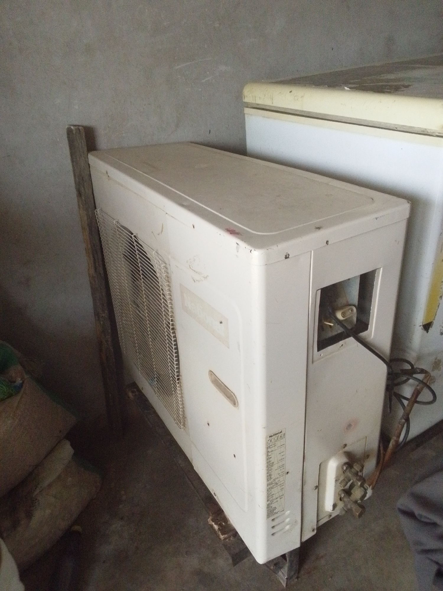 The image depicts an outdoor unit of an air conditioning system, likely an AC condenser, situated outdoors. The unit is white and appears to be made of metal. It is positioned on a wooden platform, leaning against a rough, grey concrete wall. To the left of the AC unit, a piece of rough-hewn wood is propped up against it. Behind the AC unit and to its right, the top of a white appliance, possibly a freezer or another refrigerator, is visible. The ground beneath the AC unit is a concrete floor. The scene appears to be an outdoor storage area or utility space. The overall impression is one of disuse or storage of appliances. This scene could be located in Jalingo, Nigeria, given the context provided.