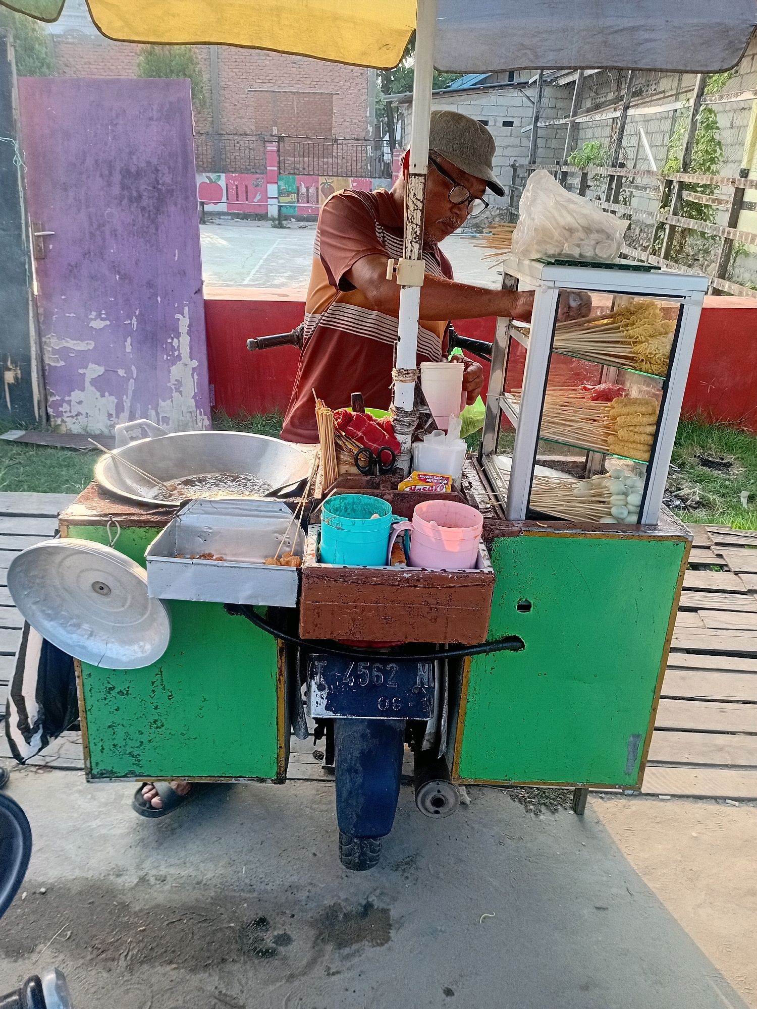 A man wearing a hat and glasses stands behind a street food cart in Anggana, Indonesia. The cart is decorated with green panels and has a yellow umbrella providing shade. The man is in the process of preparing food, with skewers of various ingredients, including what appear to be meatballs and noodles, displayed in a glass-fronted cabinet. A large wok filled with bubbling oil sits on one side of the cart, suggesting he is frying food. A motorcycle is integrated into the base of the cart, indicated by the license plate visible. The surroundings show a simple outdoor setting with grassy areas and a wooden walkway. The overall scene depicts a typical street vendor operation in a Southeast Asian urban environment.