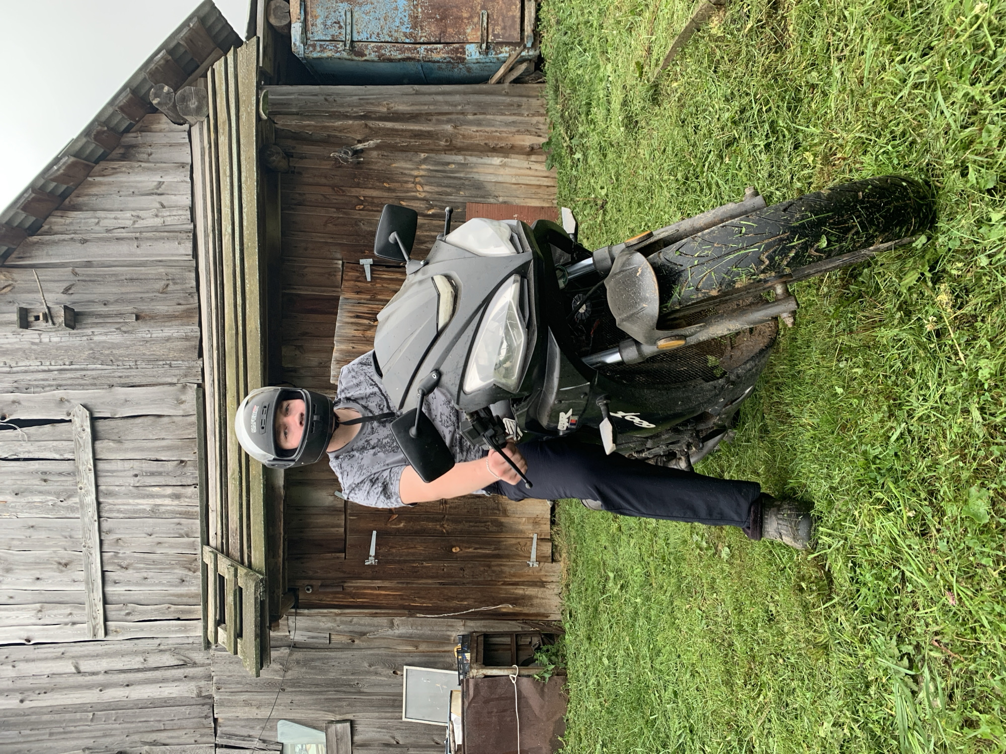 In the village of Panteleevka, Russia, a young male wearing a grey patterned t-shirt and black pants sits on a black motorcycle. He is wearing a black motorcycle helmet with grey accents. The motorcycle is parked on a grassy area in front of a weathered wooden barn. The barn appears old with a corrugated metal roof. To the left of the barn, there is a metal container and a shelf with various items. The ground is covered in green grass and some scattered debris. The motorcycle has a sleek design, with visible headlights and mirrors. The front tire of the motorcycle is covered in mud and grass.