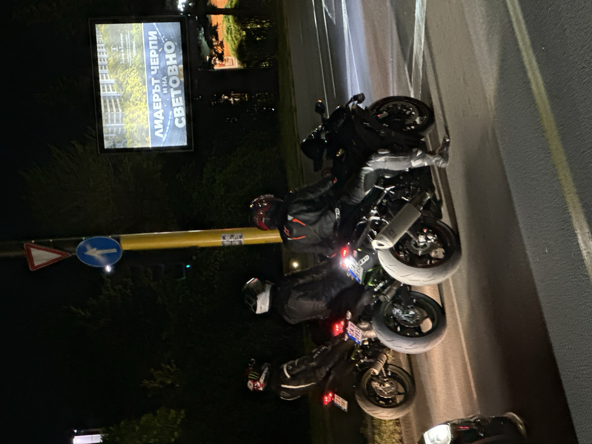 The image shows three motorcyclists stopped at a traffic light at night in an urban setting. The motorcyclists are wearing full protective gear, including helmets and leather suits. Their motorcycles are sportbikes. Behind them, there is a large illuminated billboard. The scene appears to be on a road with traffic lights, street signs, and trees visible. The lighting suggests it is nighttime and the area is illuminated by artificial lights and the billboard.