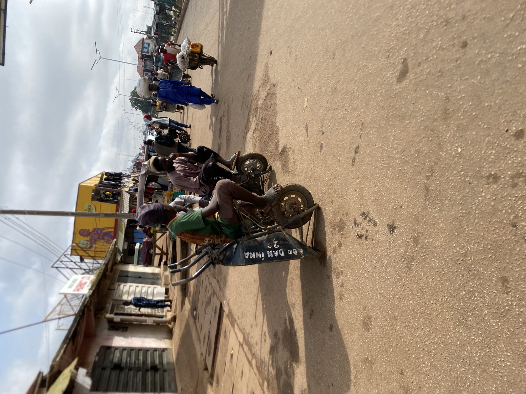 This image depicts a bustling street scene in Bukuru, Nigeria, during the daytime under a partly cloudy sky. The unpaved or gravel road is active with people and custom-made cargo vehicles.
In the foreground, two individuals are seated on distinct tricycle-like carriers, which appear to be modified for transporting goods or passengers. The vehicle closest to the viewer features "ADO DAN INNA" and the number "2" written in white on its black front panel. Several other people are visible along the street; notable figures include a person in a blue garment and another in a red shirt, both balancing large bags on their heads as they walk.
The street is flanked by various buildings, some serving as shops or residences, including one with a prominent yellow billboard displaying graphics and some unreadable text in the background. Modern streetlights line the thoroughfare, extending into the distance. The overall scene captures the vibrant daily life and commerce of a busy street, with clear shadows indicating bright sunlight despite the cloud cover.