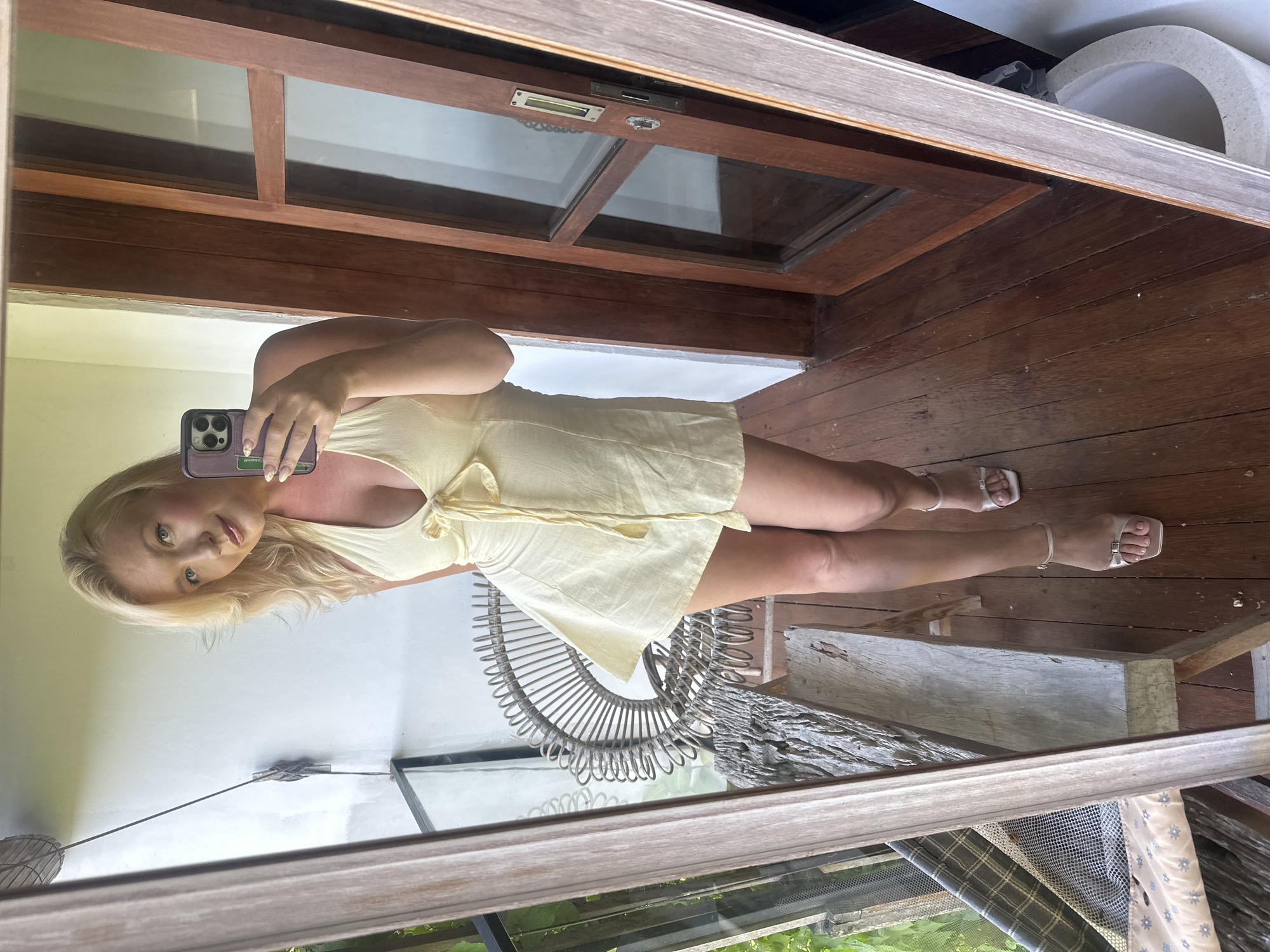 A blonde woman in a yellow mini-dress and strappy sandals stands on a wooden balcony, taking a selfie in a large mirror. She is holding a smartphone with a purple case. The balcony overlooks lush greenery, with a wicker chair visible in the reflection. A wooden door with glass panes is also present in the scene. The woman appears to be in North Kuta, Indonesia, given the tropical foliage.