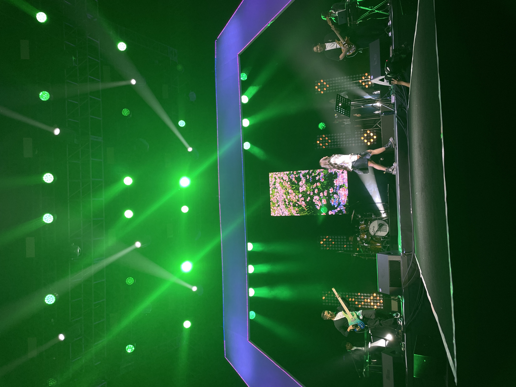 The image shows a stage setup with a performer lying on the stage floor, possibly singing or performing. There are band members on either side of the performer, playing instruments. A screen with a floral design is visible behind the performer. The stage is brightly lit with green stage lights, and there's a geometric frame around the central stage area, illuminated with blue and purple lights. Equipment like drums and speakers is also visible. The image was taken at Salembaran, Indonesia.