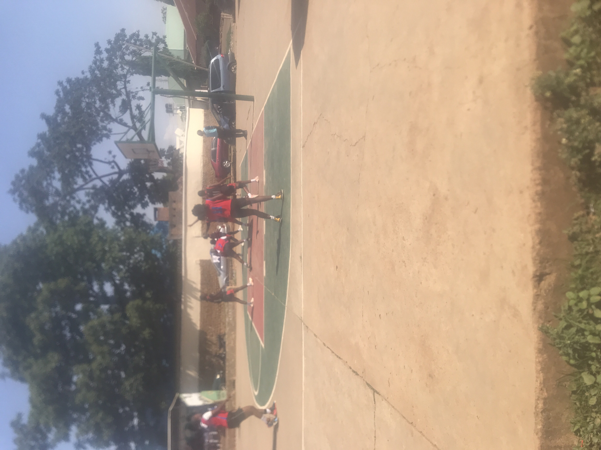 A basketball court is the primary setting. Several individuals, likely athletes, are on the court wearing sports attire. In the background, there are trees, buildings, and parked vehicles. The image was taken in Jos, Nigeria.